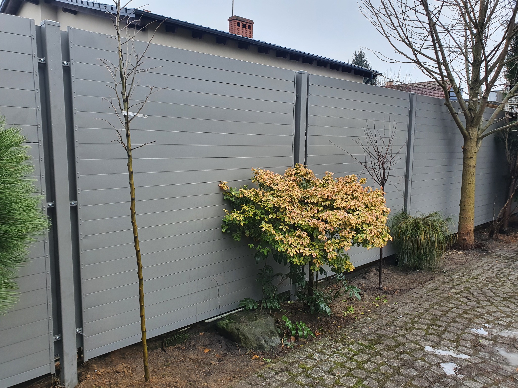 Nowoczesne ogrodzenie panelowe w kolorze szarym z poziomymi panelami, widoczne rośliny ozdobne i drzewa wzdłuż ogrodzenia, kostka brukowa na pierwszym planie, dom w tle.