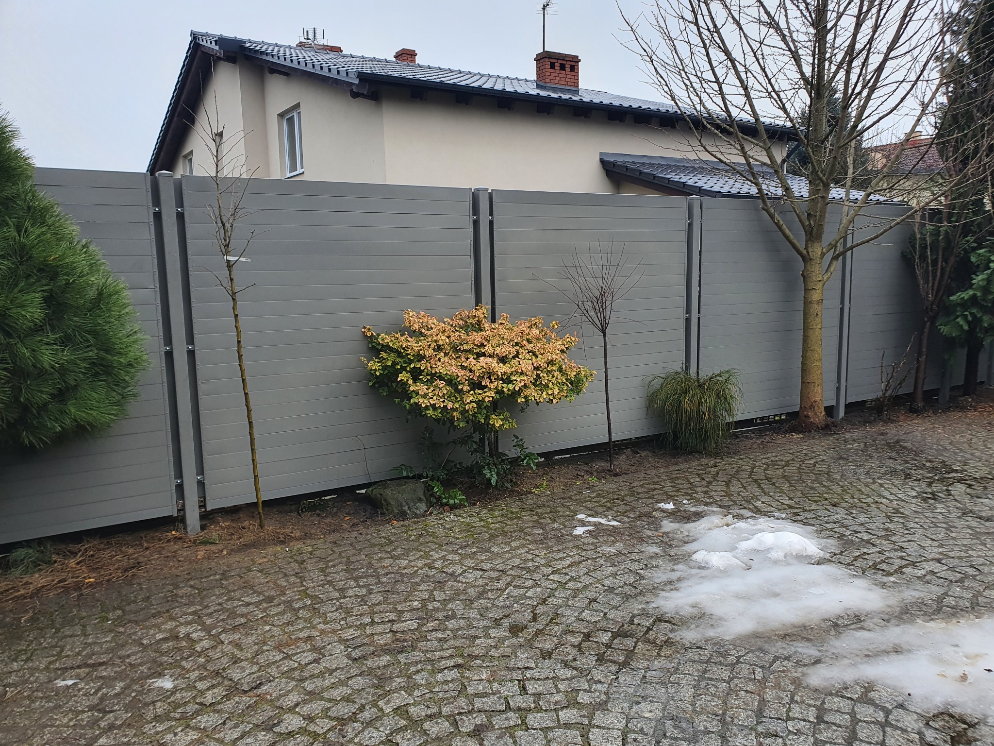 Nowoczesne ogrodzenie panelowe w kolorze szarym z poziomych desek, oddzielające posesję z kostką brukową i roślinnością od domu z ciemną dachówką.