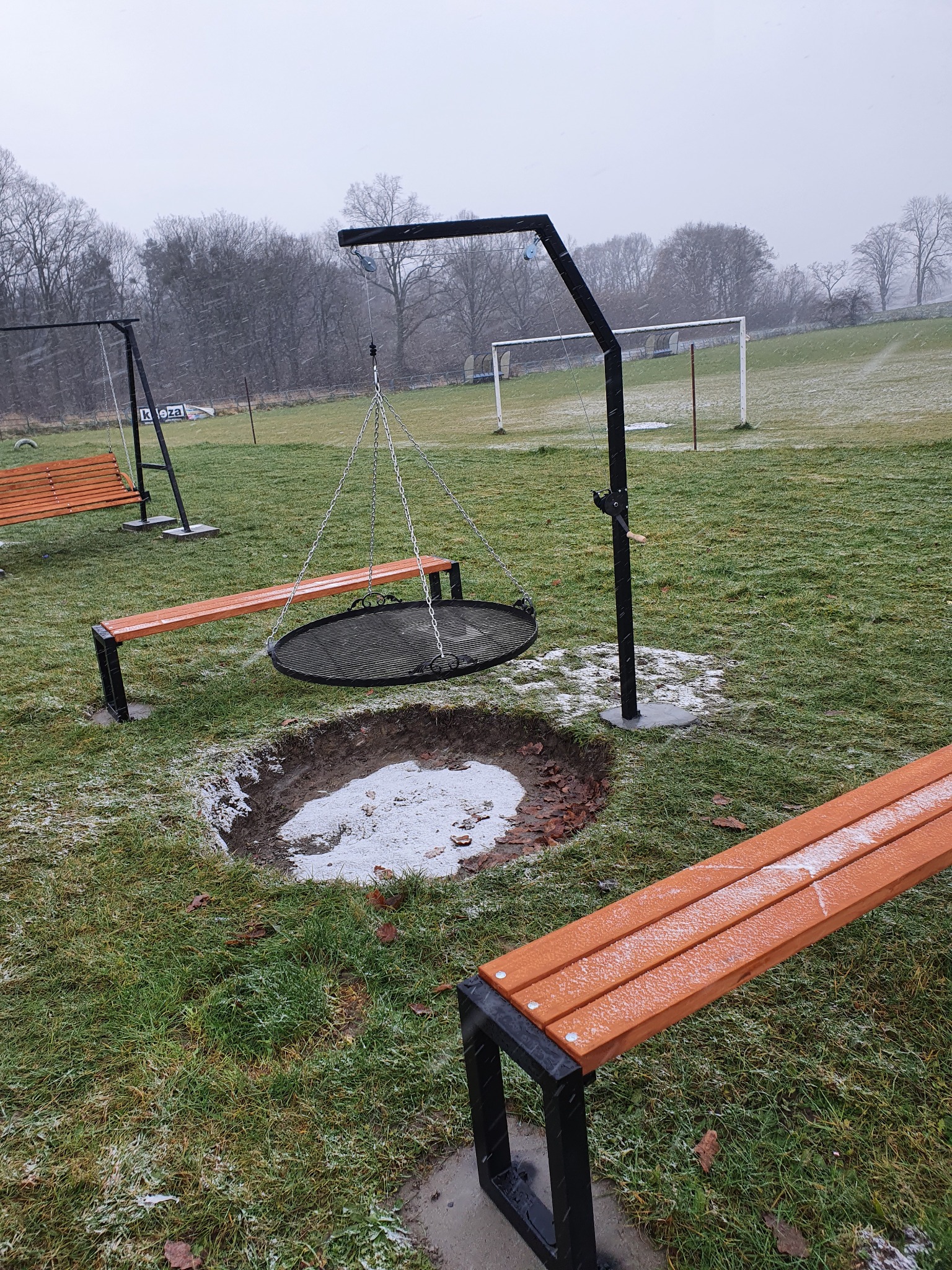 Miejsce do grillowania z metalową konstrukcją i rusztem zawieszonym na łańcuchach, otoczone drewnianymi ławkami na metalowych nogach, na trawniku z widocznym śniegiem i drzewami w tle.
