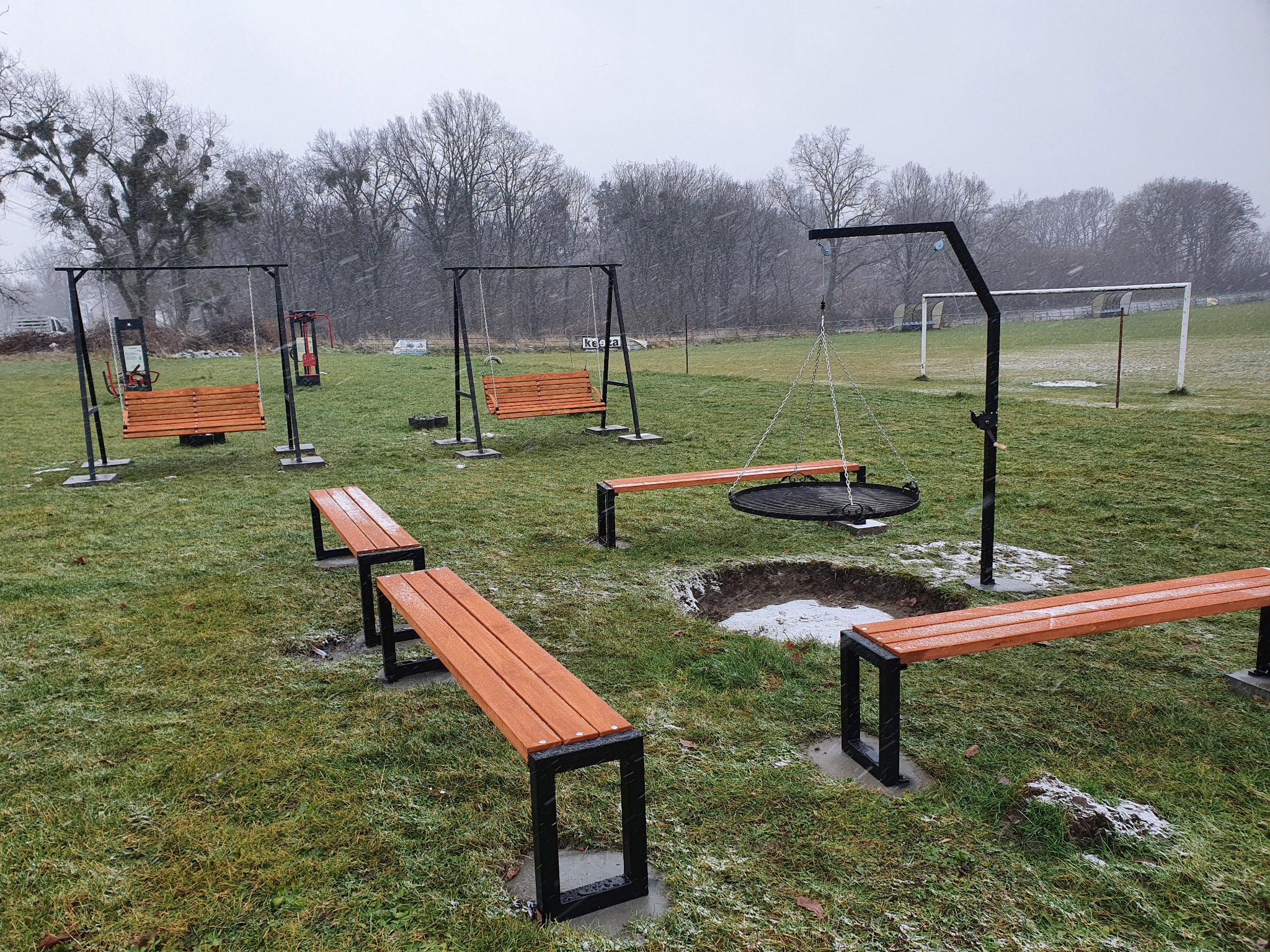 Plac zabaw na zewnątrz z huśtawkami ławkowymi z drewna i metalu, ławkami parkowymi i grillem na trójnogu, wszystko w zimowej scenerii z padającym śniegiem.