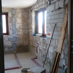 Pasternik - Surowy stan łazienki w trakcie prac: widoczne mury z cegły, instalacja wodna, prowizoryczne okablowanie elektryczne, wiadro z zaprawą i drewniany stołek.