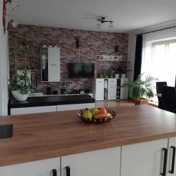 Pasternik - Jasne, przestronne wnętrze salonu z elementami dekoracyjnymi: ściana wyłożona imitacją cegły, białe meble, rośliny doniczkowe i drewniany blat wyspy kuchennej z misą owoców.