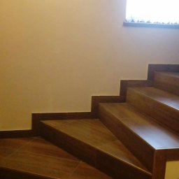 Pasternik - Wykończone schody wewnętrzne z brązowych płytek imitujących drewno, z kontrastującą ciemną listwą przypodłogową, oświetlone światłem dziennym z okna na ścianie.