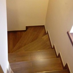 Pasternik - Widok z góry na schody wyłożone płytkami imitującymi drewno, z ciemnobrązowymi obrzeżami i jasnymi ścianami.