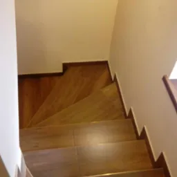 Widok z góry na schody wyłożone płytkami imitującymi drewno, z ciemnobrązowymi obrzeżami i jasnymi ścianami.