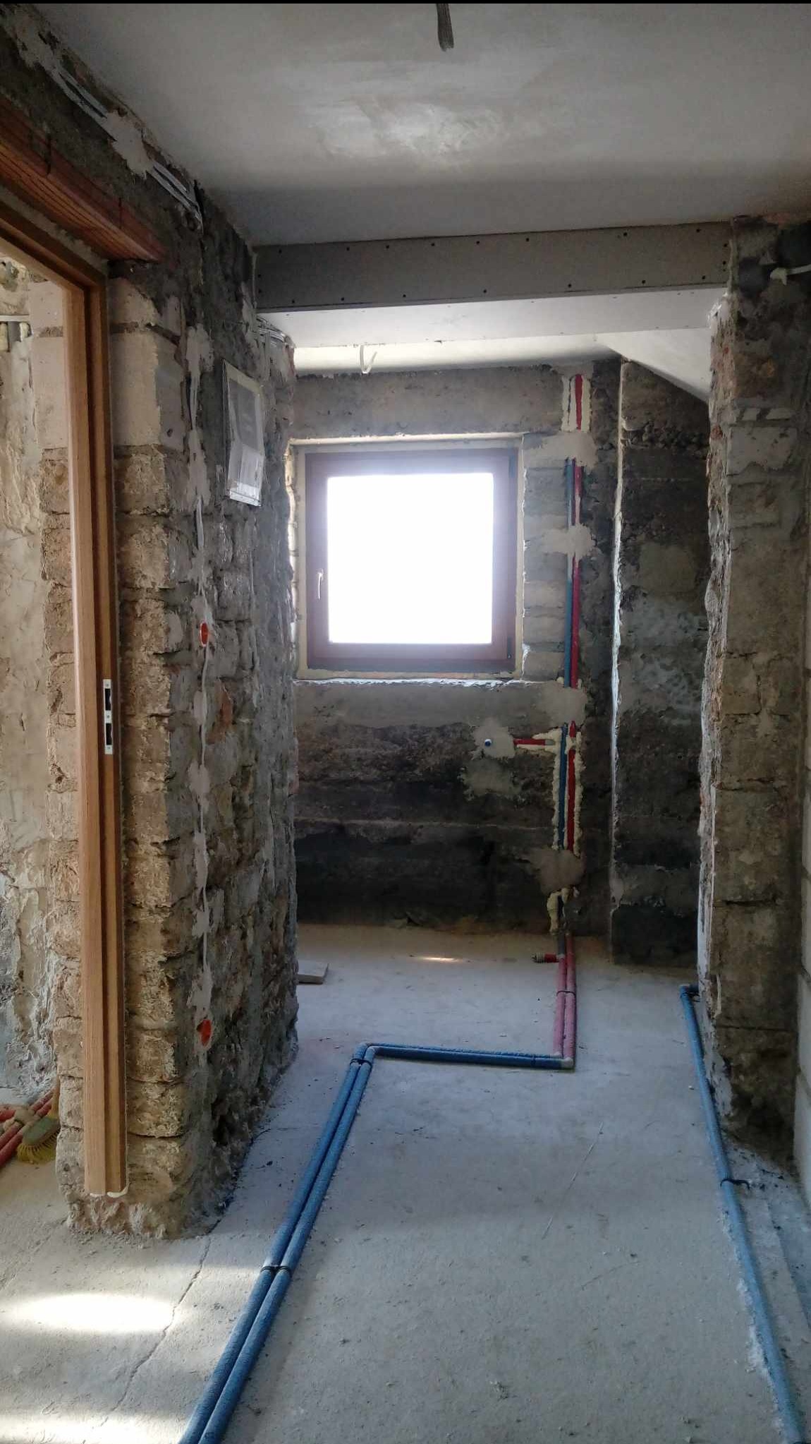 Wnętrze w trakcie prac przygotowawczych do wykończenia: surowe ściany z cegły i betonu, nowe okno z drewnianą ramą, instalacja wodna w kolorze czerwonym i niebieskim na ścianie, rury instalacyjne...