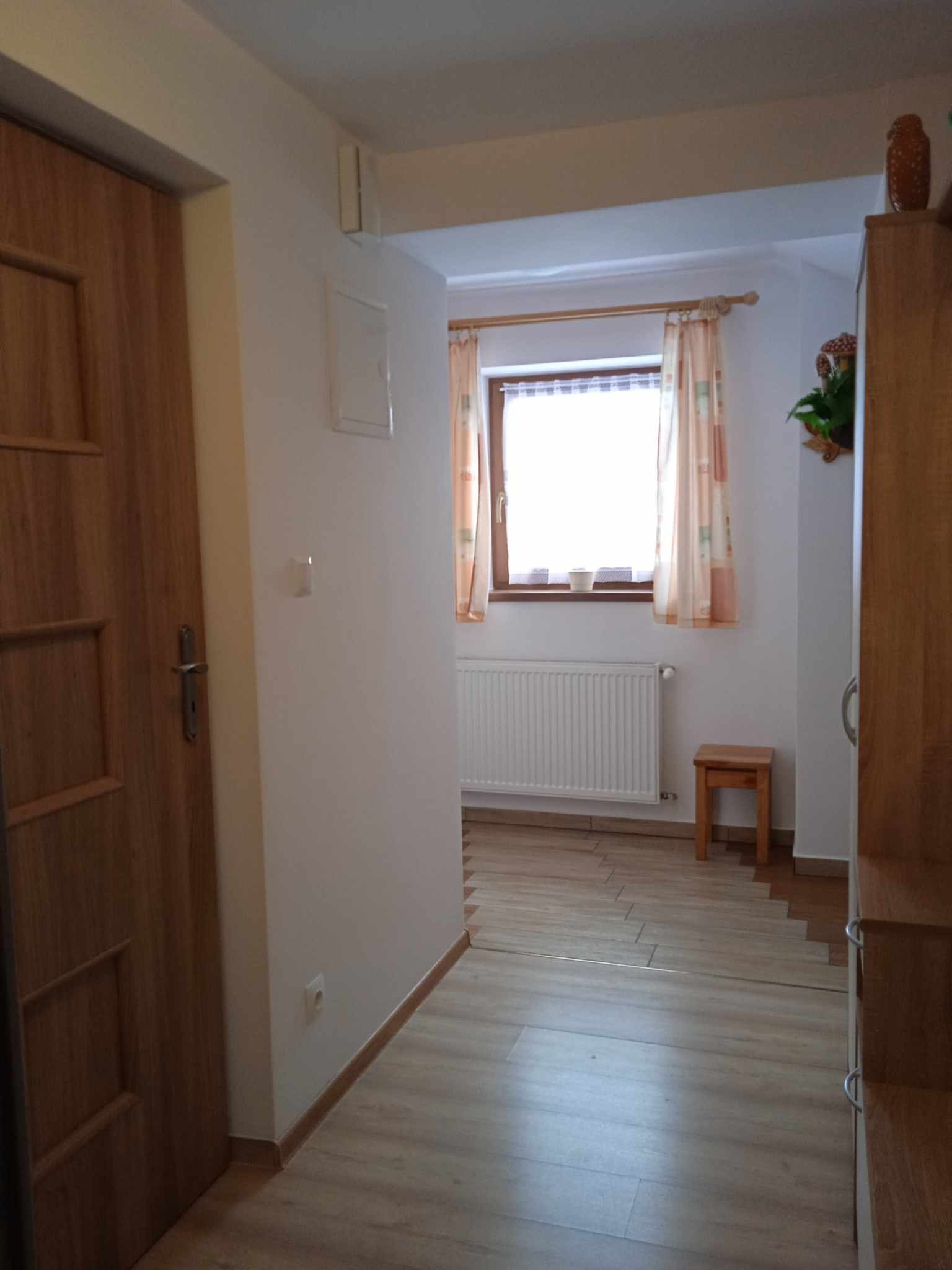 Wnętrze korytarza po remoncie z drewnianą podłogą, drzwiami i szafą, widoczny grzejnik pod oknem z zasłonami i mały drewniany stołek.