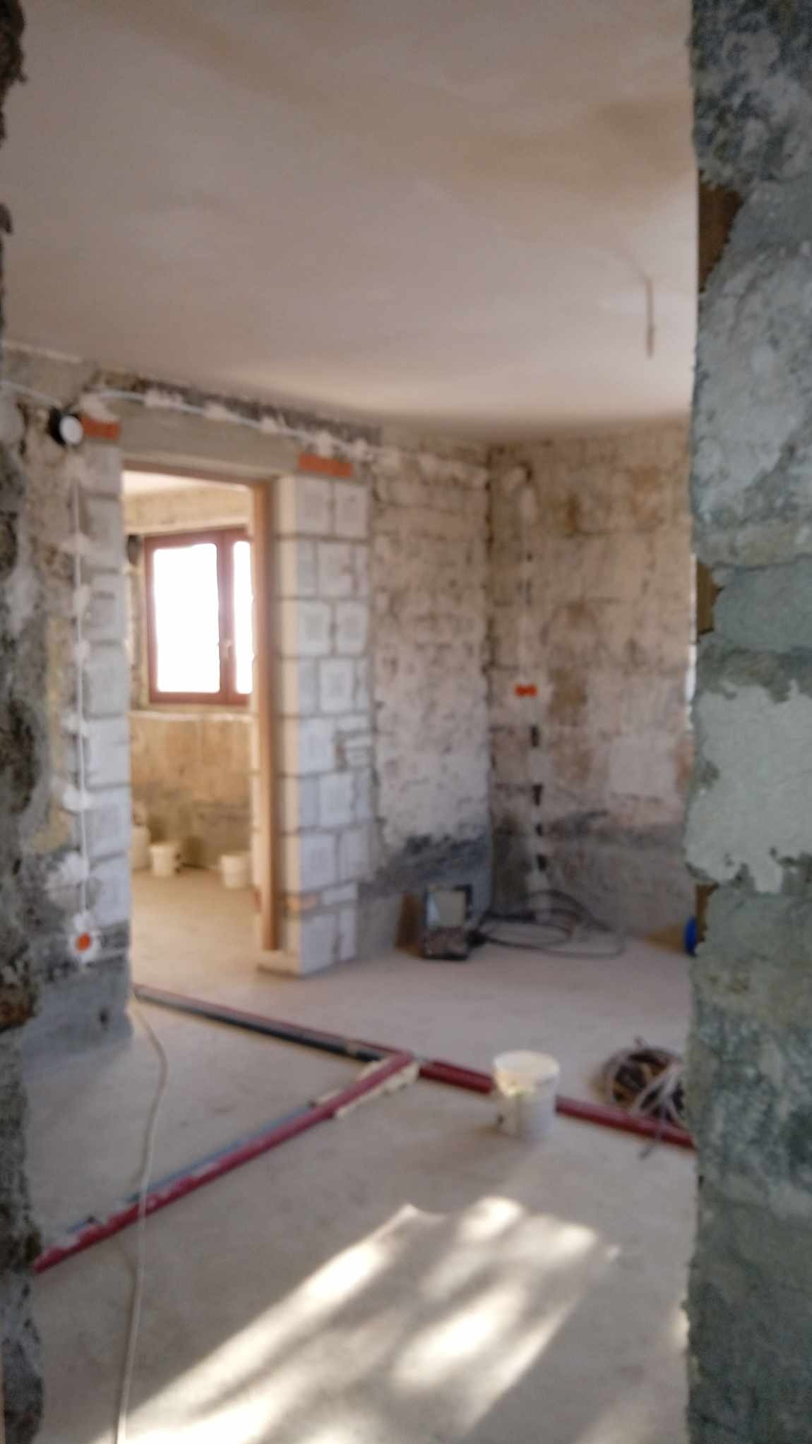 Surowy stan wnętrza w trakcie prac: ściany z bloków, widoczne instalacje elektryczne i rury na podłodze, przygotowane do dalszych prac wykończeniowych.