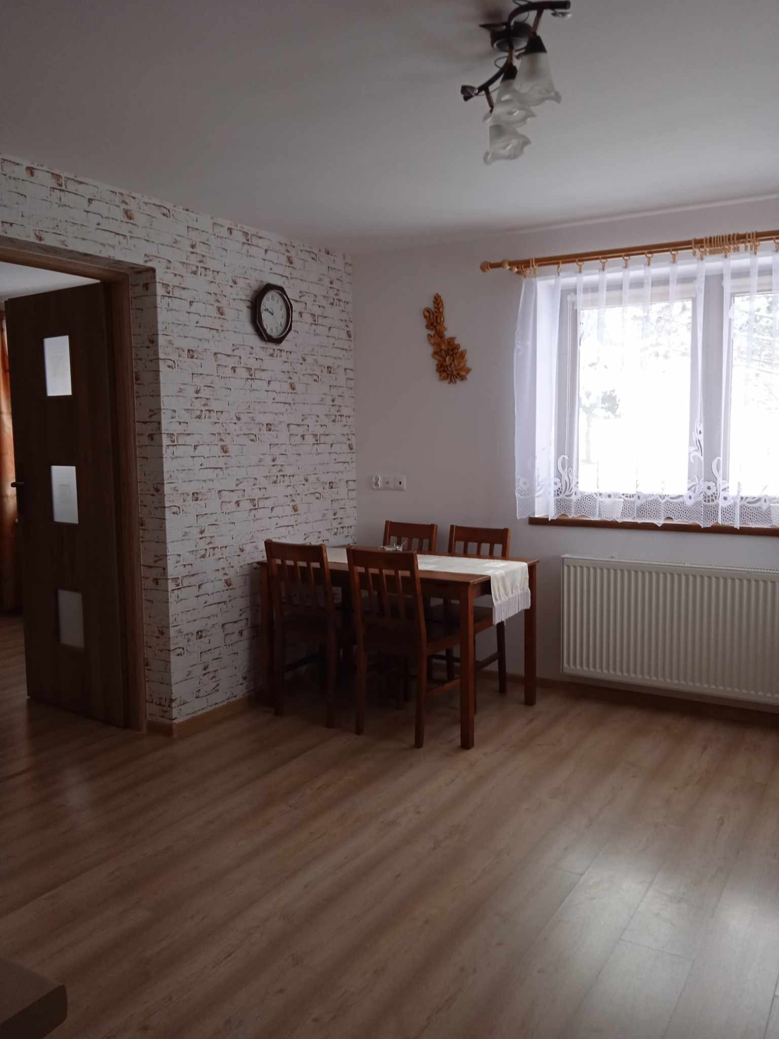 Jasne wnętrze pokoju z drewnianym stołem nakrytym białym obrusem, krzesłami, grzejnikiem pod oknem z białymi firankami i ścianą wykończoną tapetą imitującą białą cegłę, na której wisi zegar.