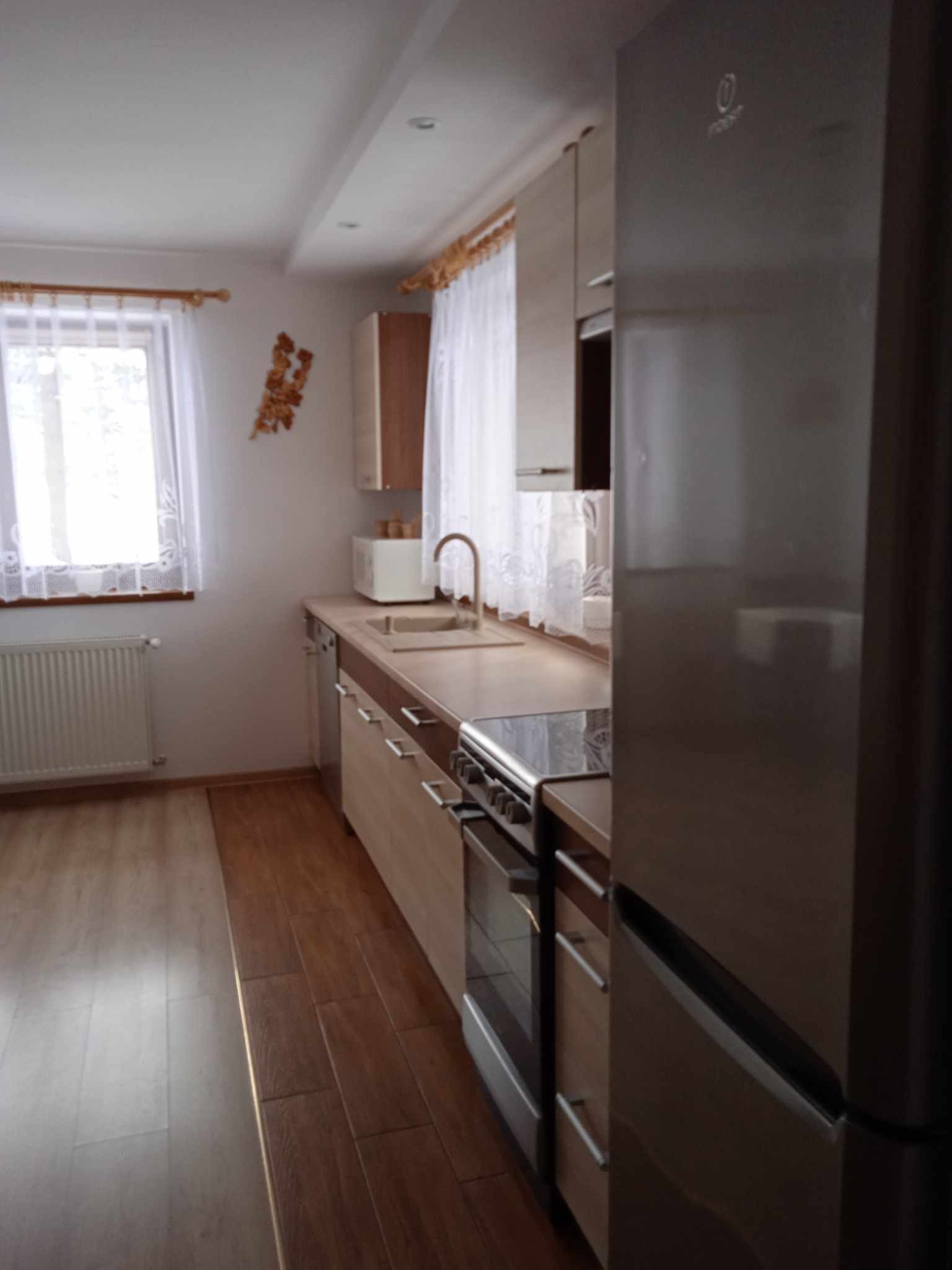 Jasna kuchnia z drewnianymi panelami podłogowymi, beżowymi szafkami i białymi zasłonami w oknie, widok na kuchenkę i lodówkę.