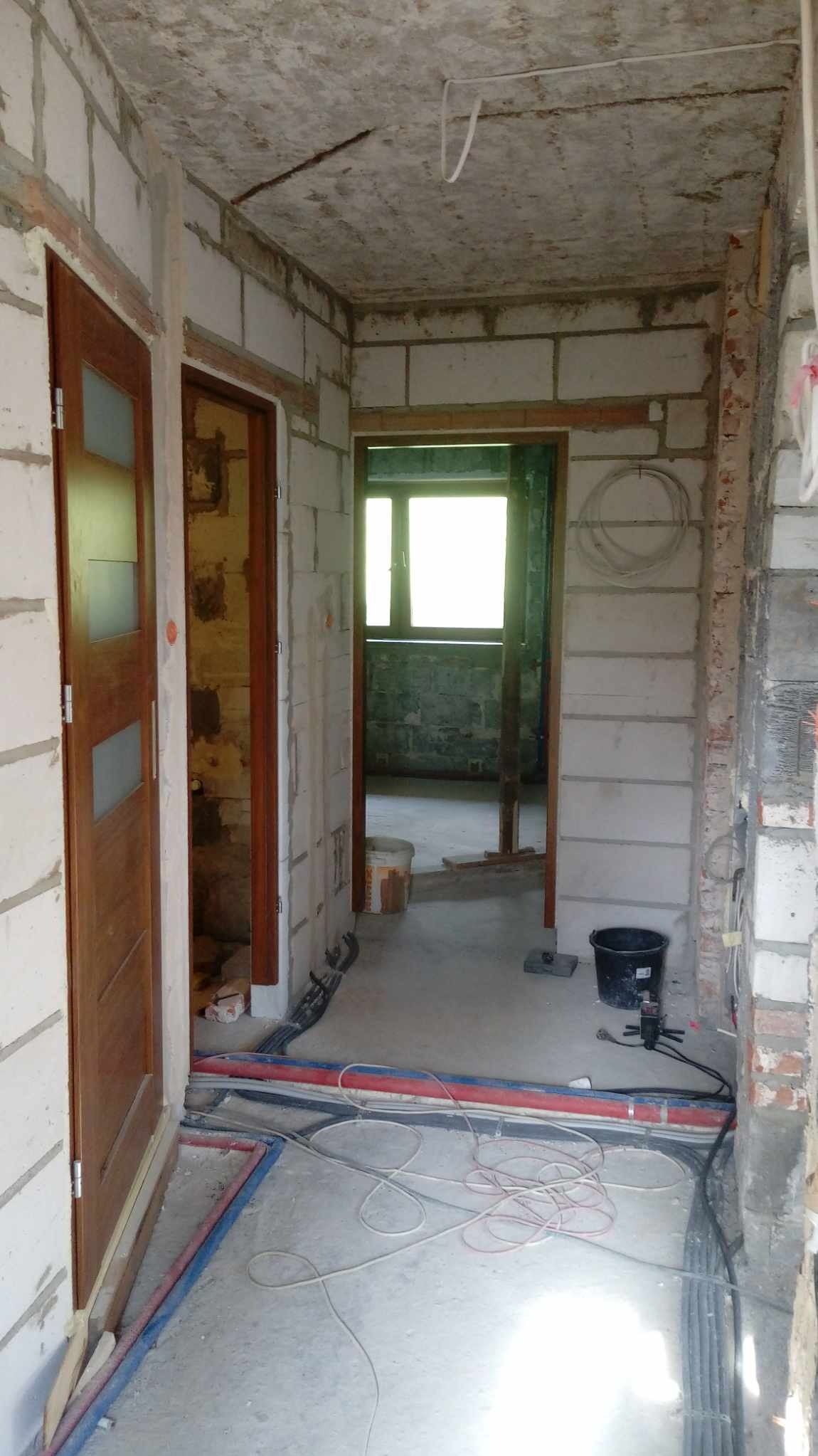 Korytarz w trakcie prac wykończeniowych: surowe ściany z betonu komórkowego, widoczne instalacje elektryczne i hydrauliczne na podłodze, drewniane ościeżnice drzwiowe, sufit w stanie surowym, widok...