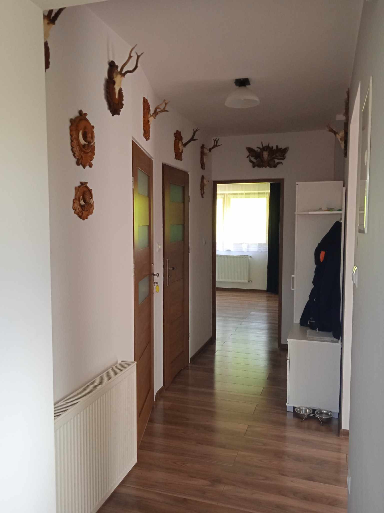 Jasny korytarz z drewnianą podłogą, białymi ścianami udekorowanymi trofeami myśliwskimi i grzejnikiem, widok na dalsze pomieszczenia.