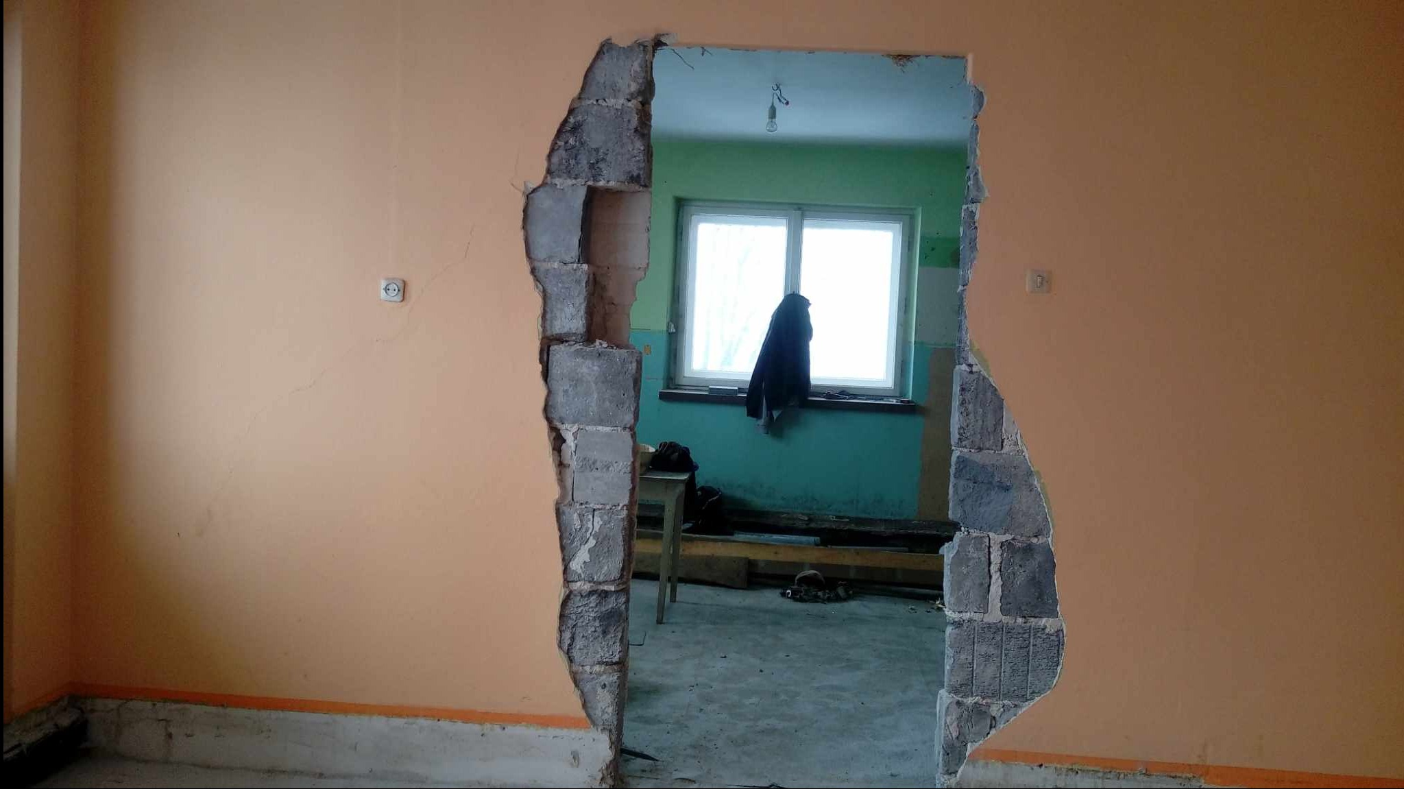 Otwór w ścianie z widocznym fragmentem instalacji elektrycznej, odsłonięte bloki betonowe, widok na pomieszczenie z oknem, ubraniem wiszącym na parapecie i prowizorycznym wyposażeniem.