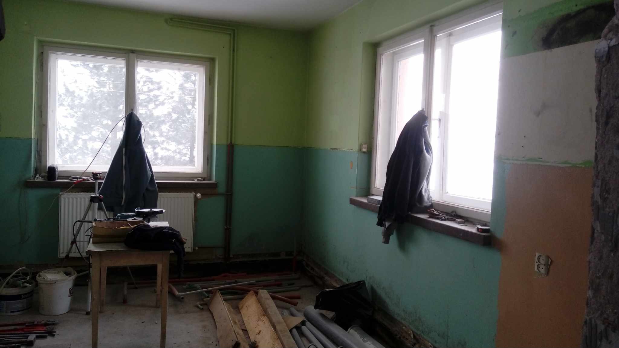 Pokój w trakcie odświeżania: zielone ściany w dwóch odcieniach, okna przesłonięte ubraniami, narzędzia na stole, widoczne rury i listwy przypodłogowe na podłodze, fragment ściany przygotowany...