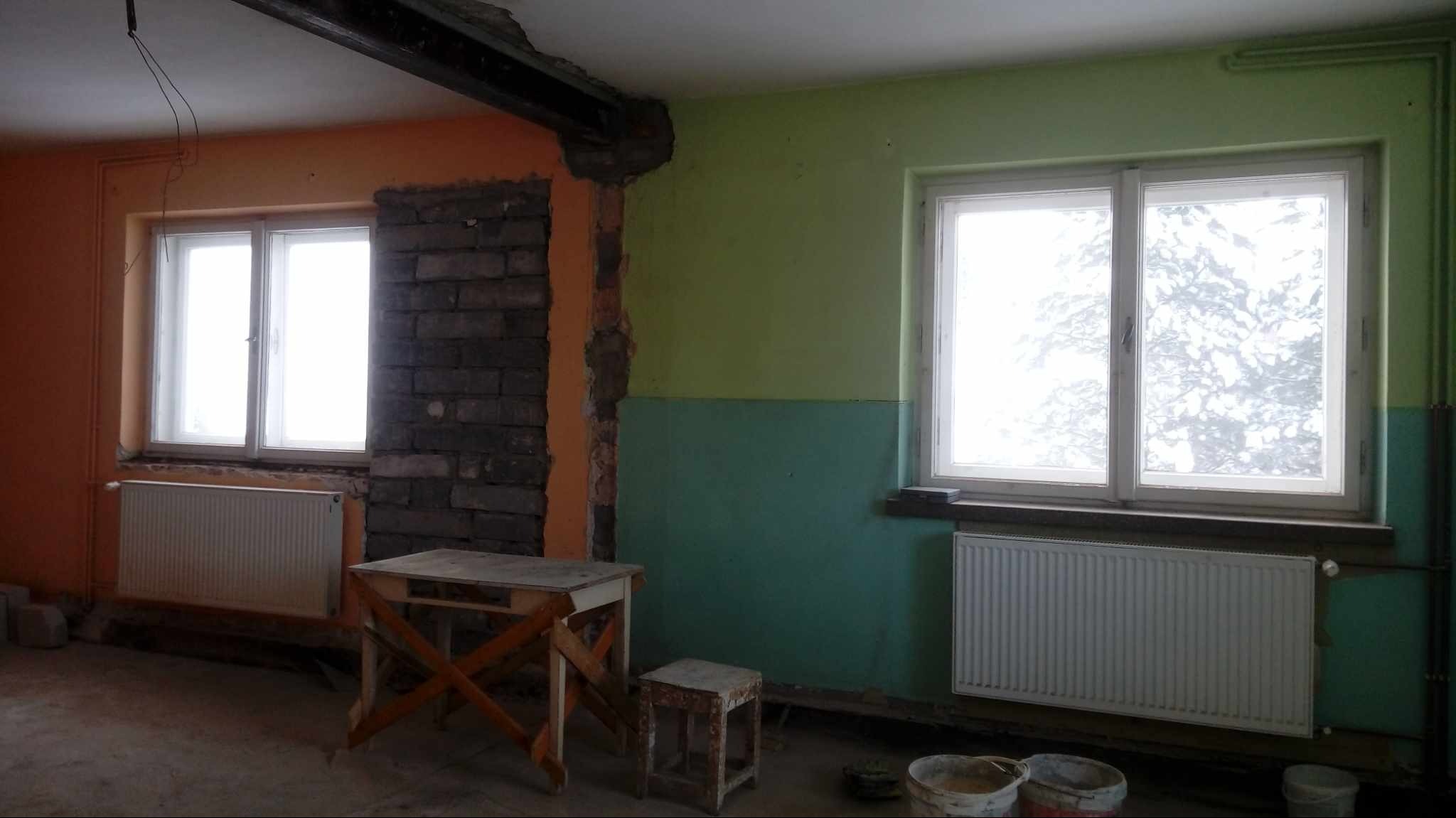 Pokój w trakcie remontu: ściany częściowo pomalowane na pomarańczowo i zielono, fragment ściany z odsłoniętą cegłą, drewniana belka stropowa, okna z grzejnikami, prowizoryczny stolik i stołek...