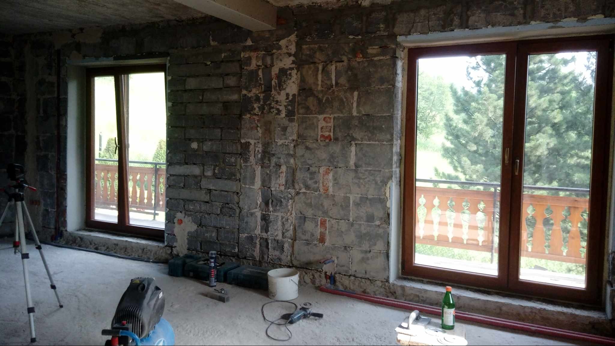 Surowy stan wnętrza z dwoma oknami przed wykończeniem, widoczne narzędzia i materiały budowlane, ściany z bloków z fragmentami starego tynku, widok na zewnątrz przez okna.