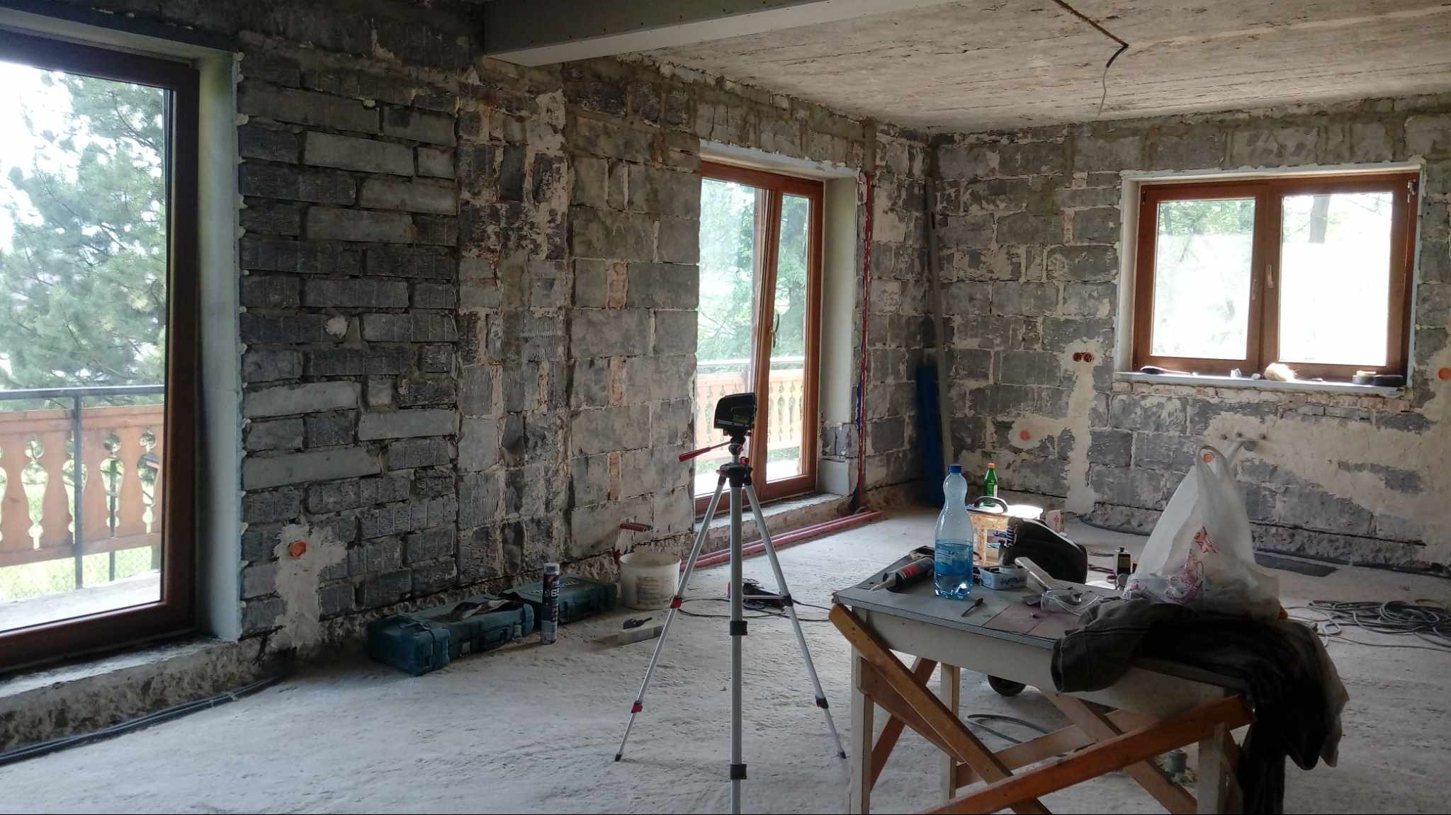 Surowe mury z widocznymi śladami po instalacjach, drewniana stolarka okienna, poziomica laserowa na statywie, prowizoryczny stół z narzędziami i butelką wody w pomieszczeniu w trakcie prac...