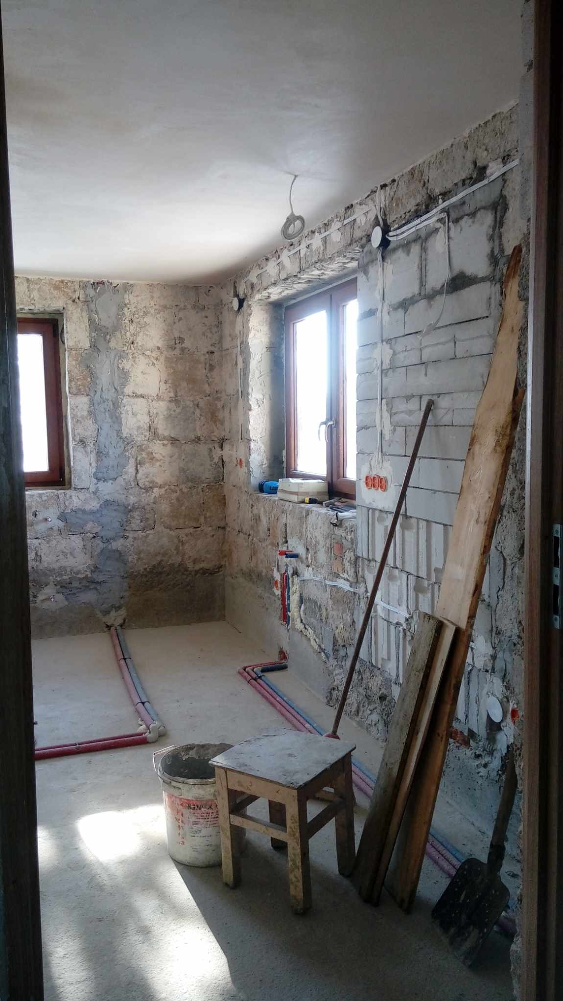 Surowy stan łazienki w trakcie prac: widoczne mury z cegły, instalacja wodna, prowizoryczne okablowanie elektryczne, wiadro z zaprawą i drewniany stołek.