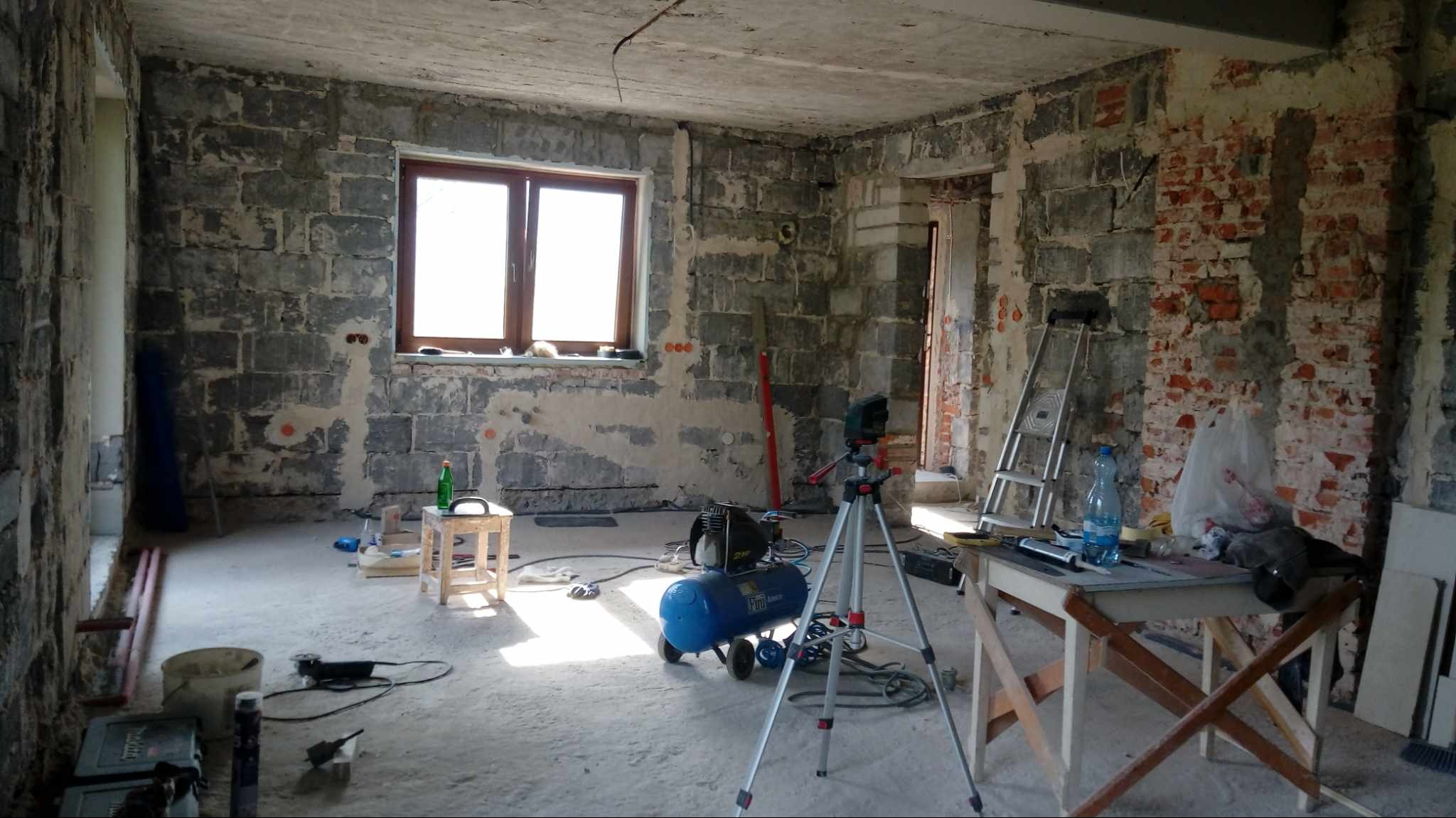 Surowe wnętrze w trakcie prac wykończeniowych: odsłonięte cegły i beton, widoczne instalacje, poziomica, kompresor, laser na statywie, drabina, prowizoryczny stół roboczy z narzędziami, butelka...