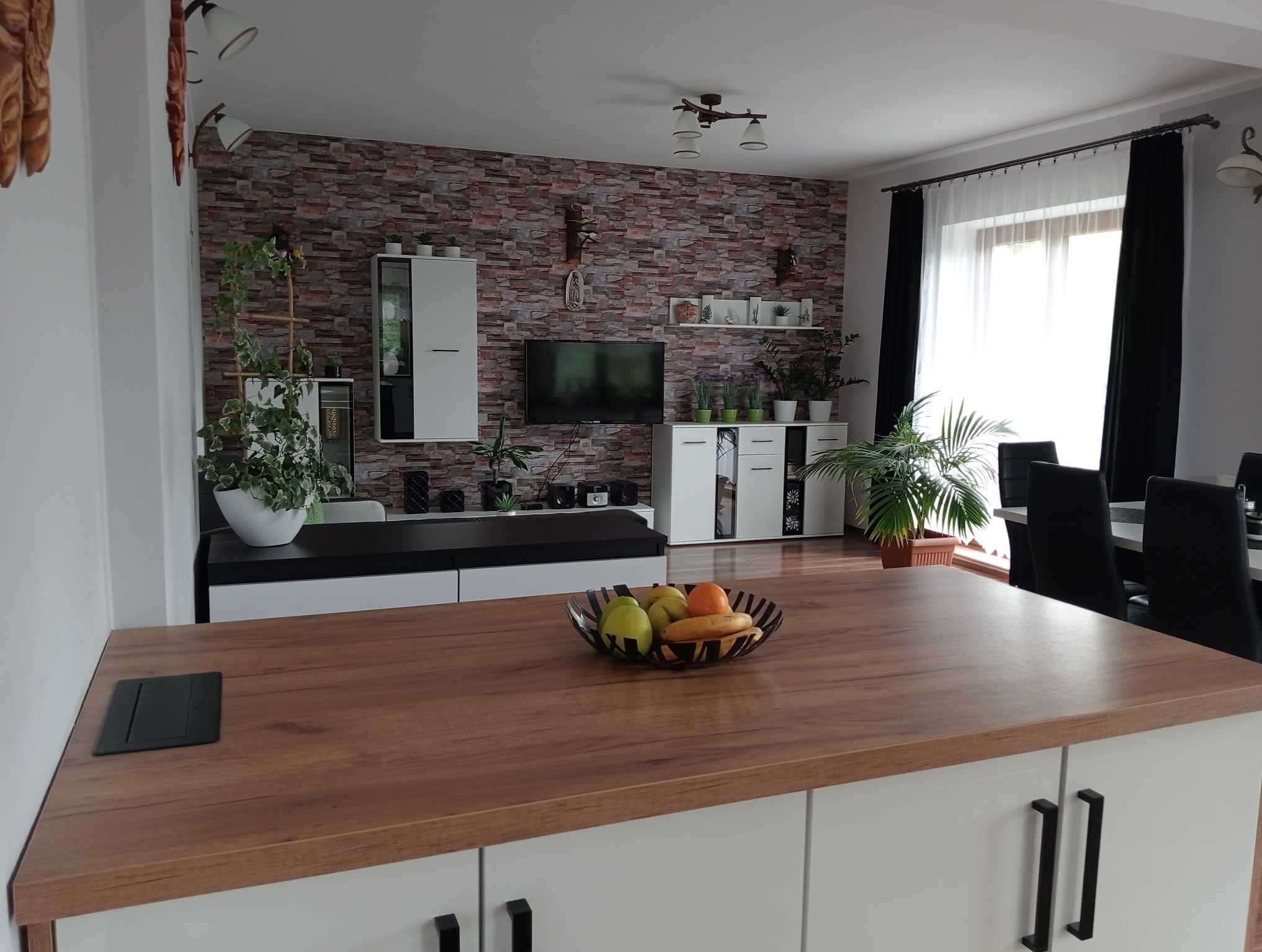 Jasne, przestronne wnętrze salonu z elementami dekoracyjnymi: ściana wyłożona imitacją cegły, białe meble, rośliny doniczkowe i drewniany blat wyspy kuchennej z misą owoców.