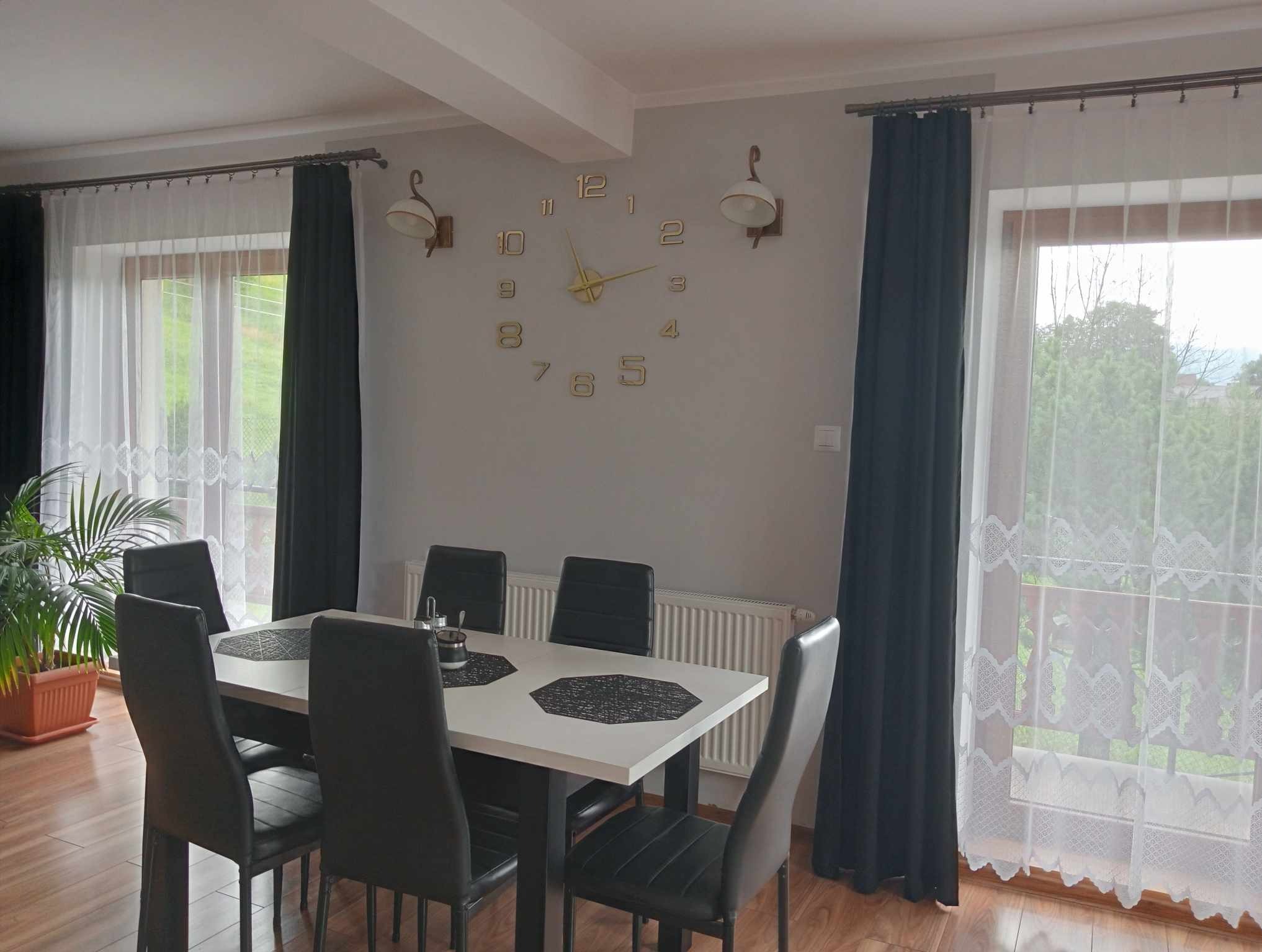 Jasne wnętrze jadalni z białym stołem, czarnymi krzesłami, dużym zegarem ściennym i dekoracyjnymi lampami ściennymi w stylu retro.