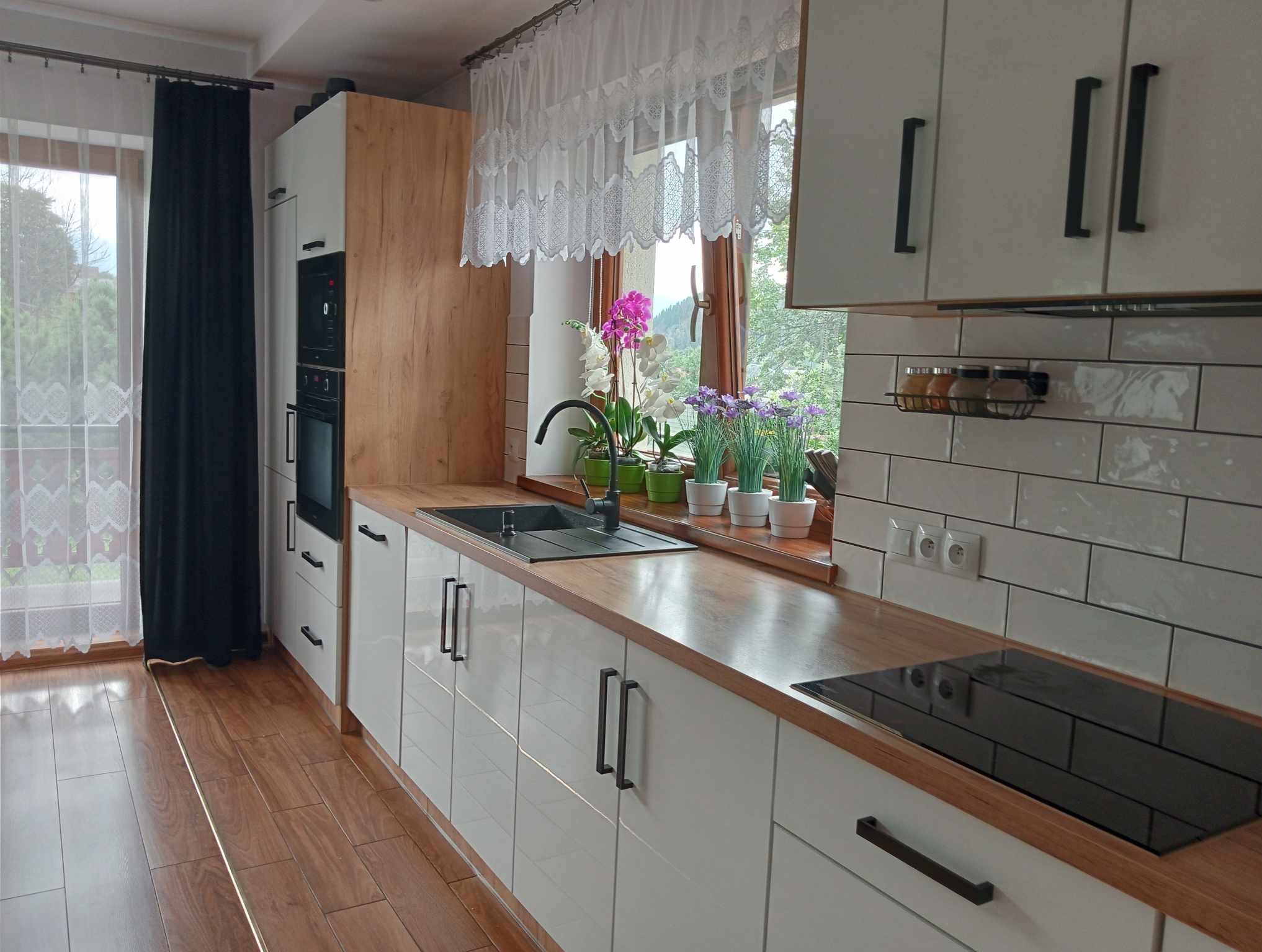 Jasna kuchnia z białymi szafkami, drewnianym blatem i czarnymi uchwytami, z widokiem na zieleń za oknem ozdobionym kwiatami w doniczkach.