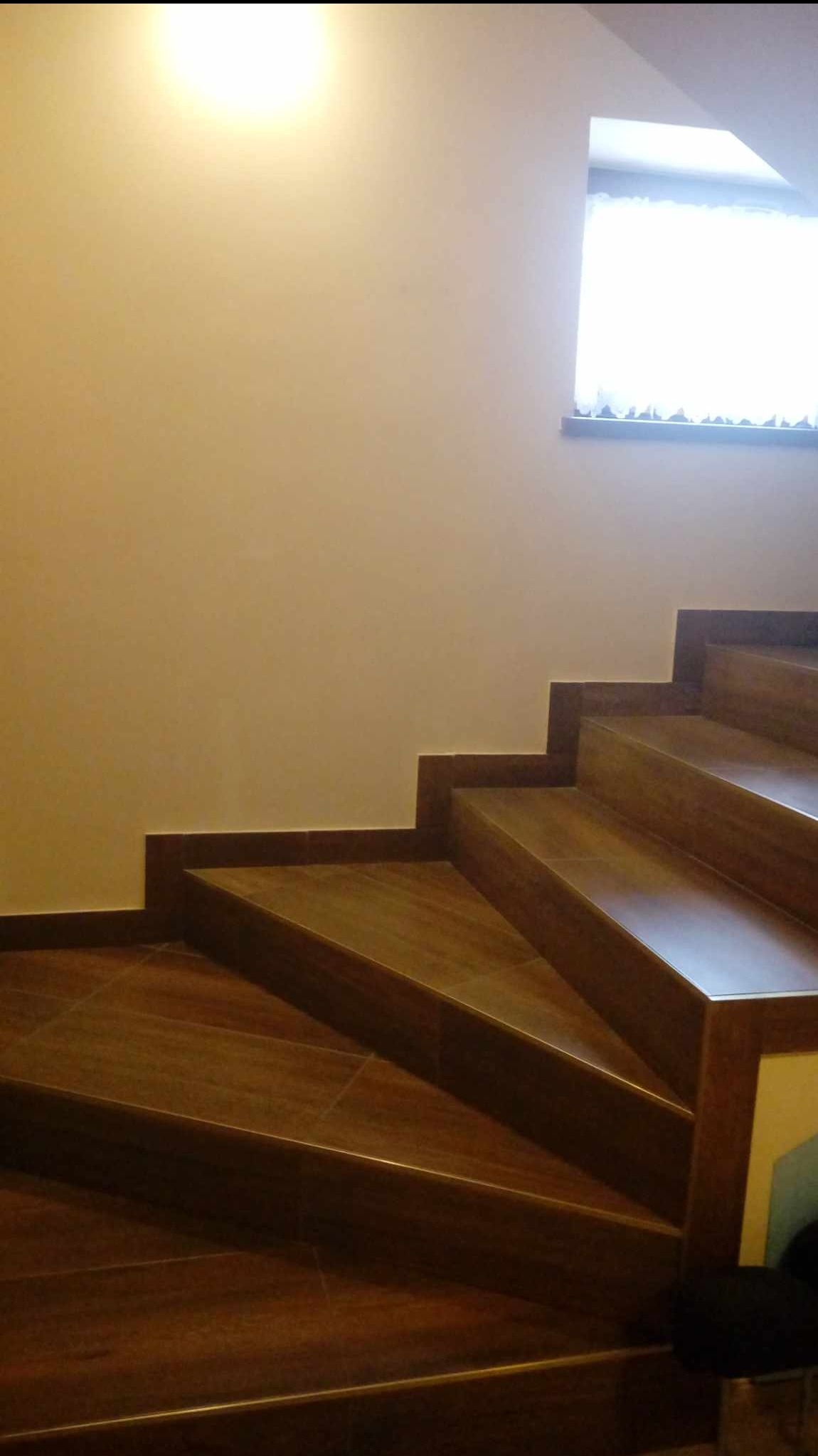 Wykończone schody wewnętrzne z brązowych płytek imitujących drewno, z kontrastującą ciemną listwą przypodłogową, oświetlone światłem dziennym z okna na ścianie.