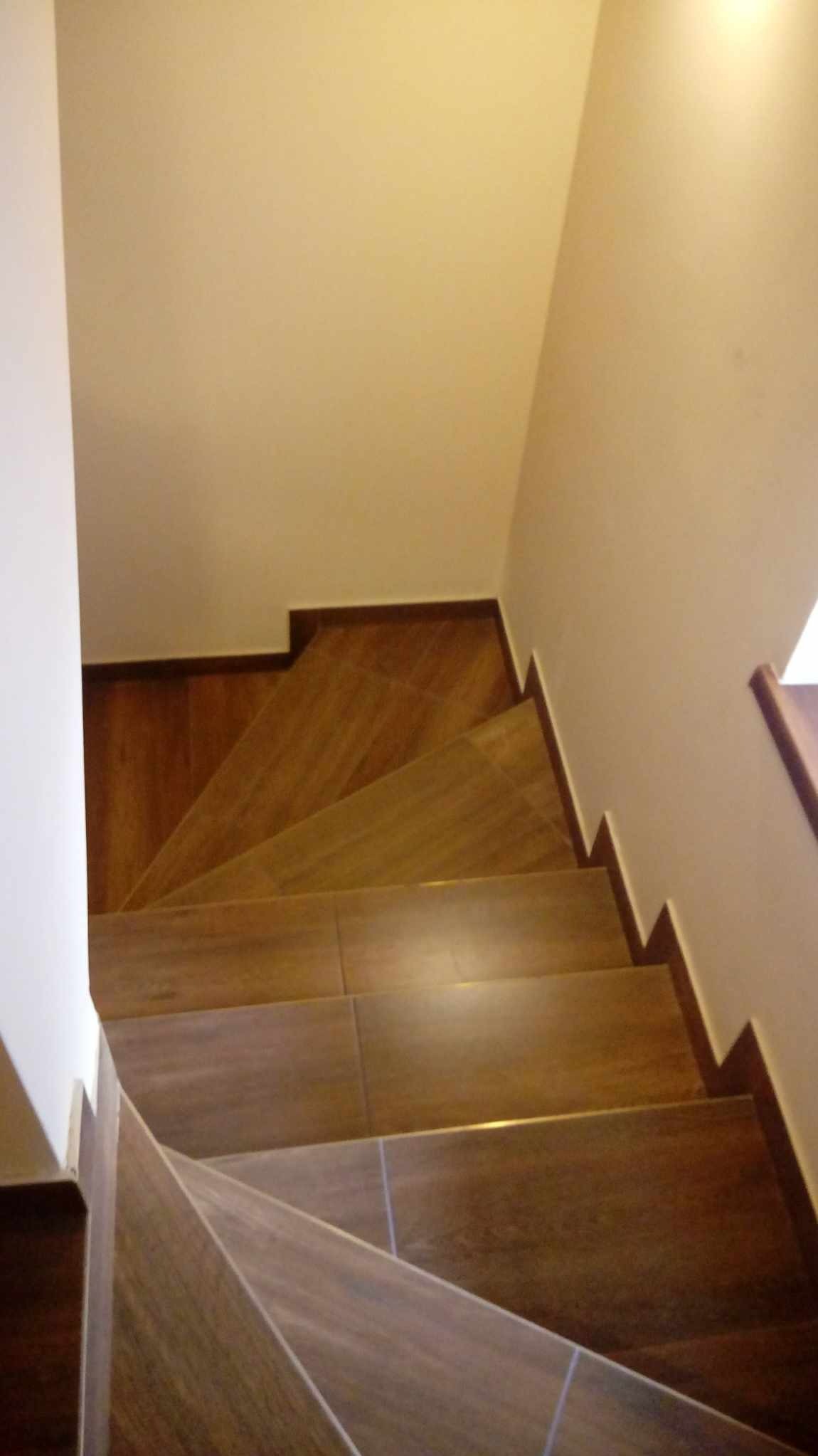 Widok z góry na schody wyłożone płytkami imitującymi drewno, z ciemnobrązowymi obrzeżami i jasnymi ścianami.