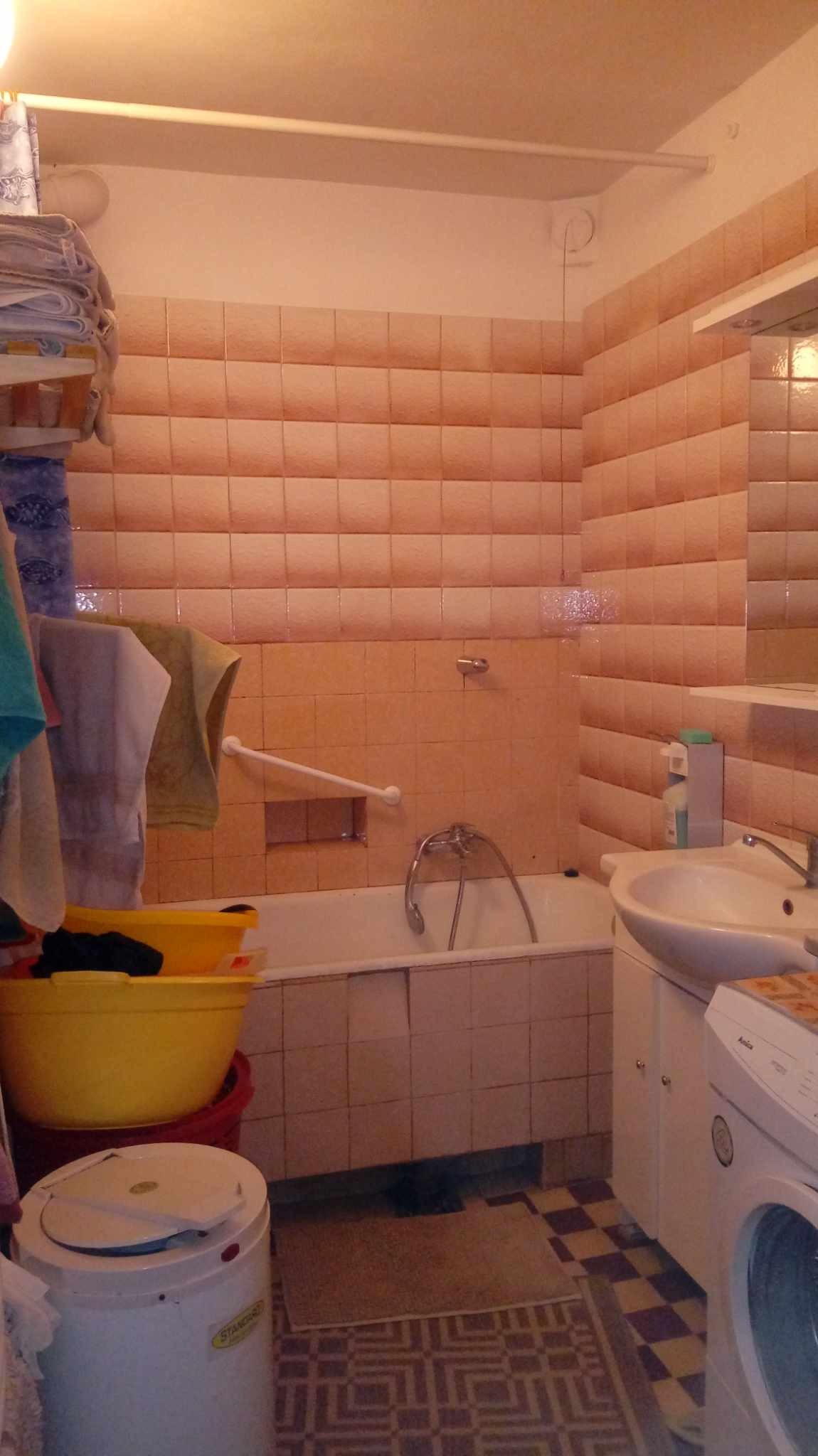 Standardowa łazienka z różowymi kafelkami, białą wanną i umywalką, pralką Amica, żółtymi miskami z praniem, oraz grzejnikiem ceramicznym.