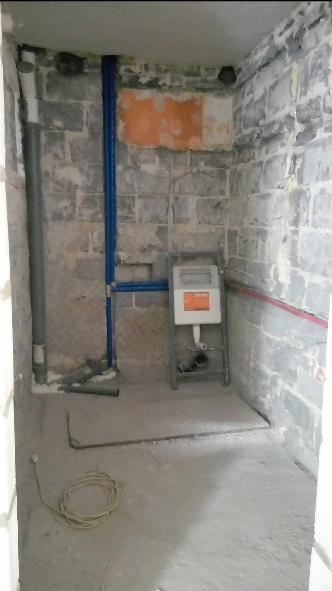 Surowa łazienka w trakcie prac: widoczne nieotynkowane ściany z cegły, niebieskie rury instalacji wodnej, szara rura kanalizacyjna, stelaż podtynkowy WC marki Pando i zwój kabla elektrycznego...