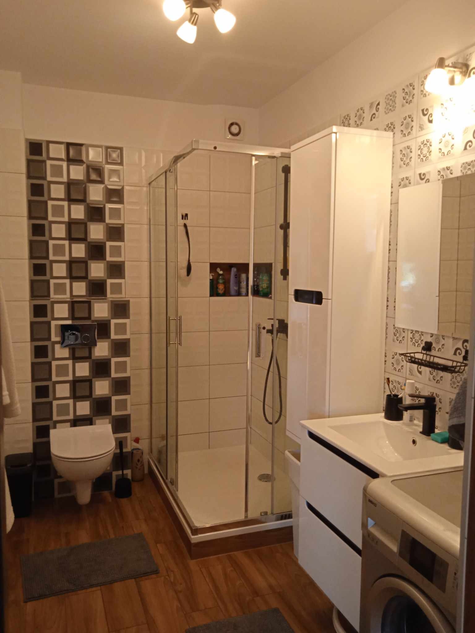 Nowoczesna łazienka z kabiną prysznicową, umywalką z szafką, pralką i dekoracyjnymi płytkami na ścianie.