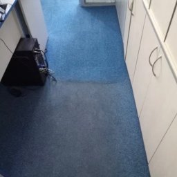 Kostiantyn Kalymaha - Fragment biura z niebieską wykładziną dywanową, widoczna różnica w odcieniu po czyszczeniu na części powierzchni, przy ścianie białe szafki z metalowymi uchwytami i biurko z komputerem.