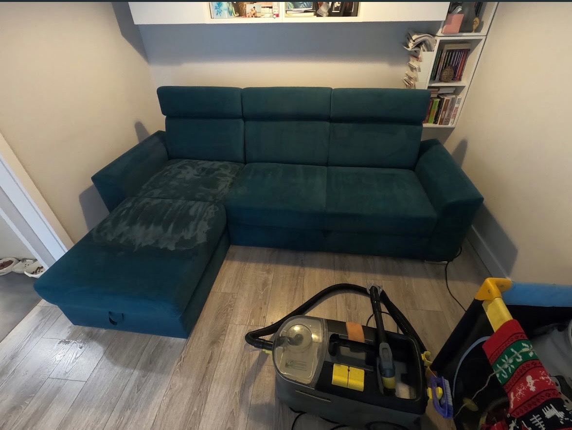 Turkusowa kanapa narożna z widoczną różnicą w czystości po lewej stronie siedziska, obok odkurzacz piorący i dziecięcy kojec.