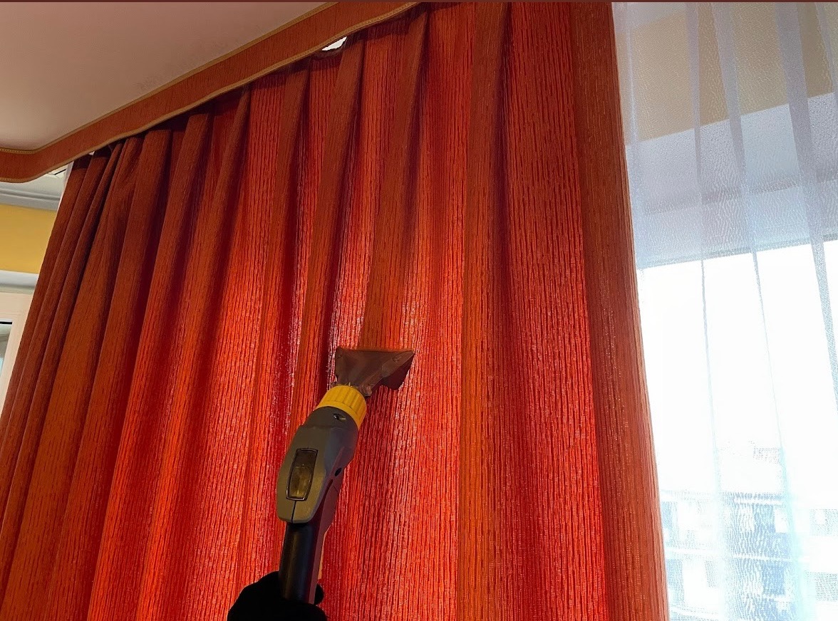 Pomarańczowe zasłony czyszczone parowo z użyciem dyszy i odkurzacza parowego, widoczne okno z firanką w tle.
