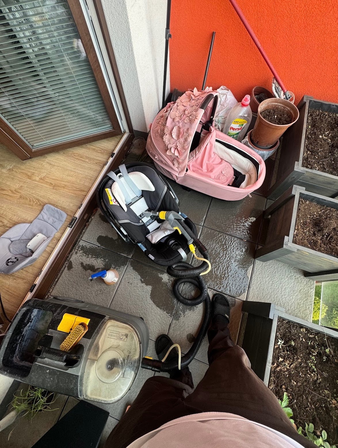 Czyszczenie fotelika samochodowego na mokro na balkonie, widoczny odkurzacz piorący, woda i akcesoria, obok wózek dziecięcy i donice z ziemią.