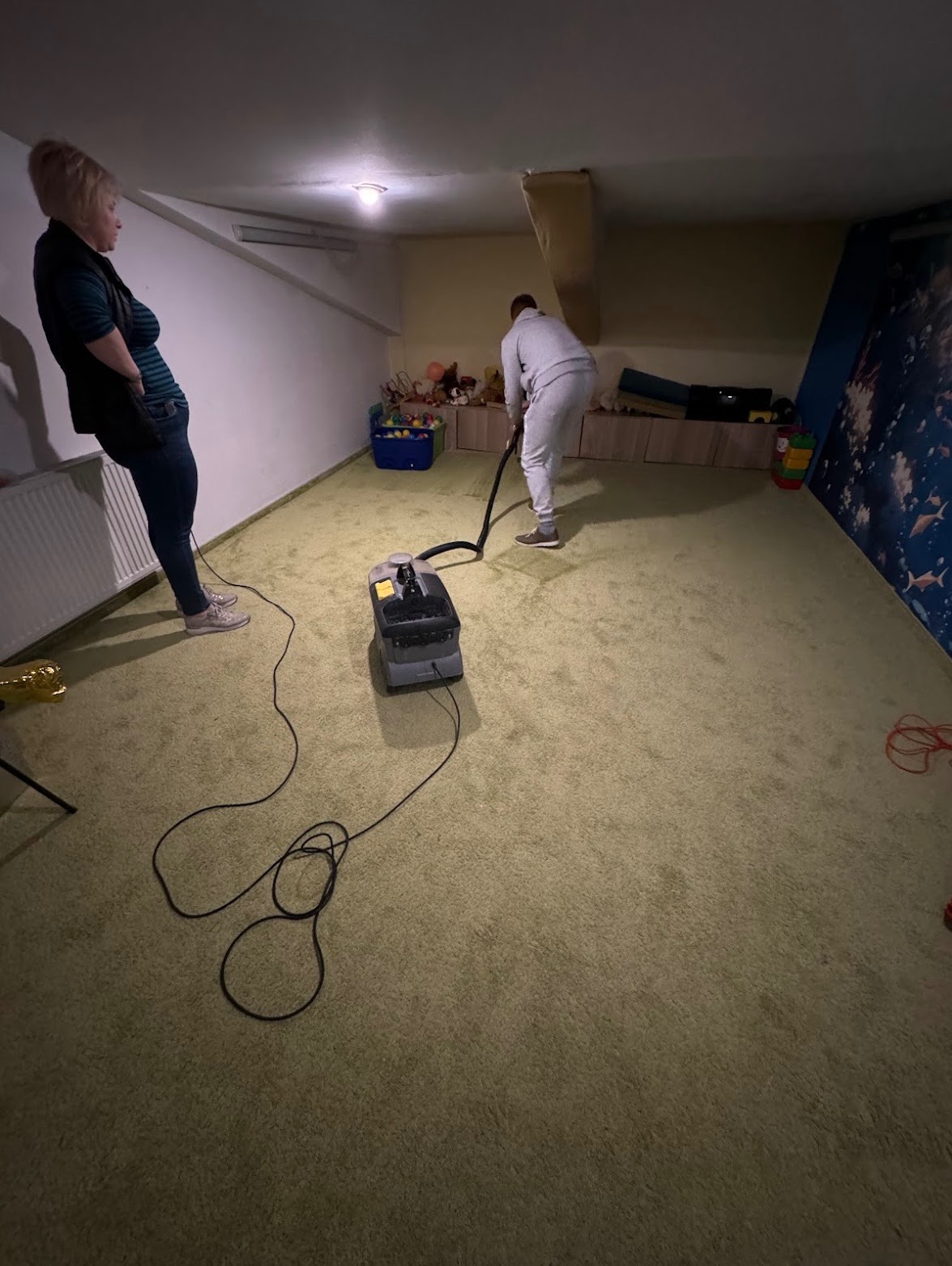 Osoba w szarym dresie czyści zielony dywan w pokoju dziecięcym za pomocą profesjonalnego odkurzacza piorącego; druga osoba nadzoruje pracę.