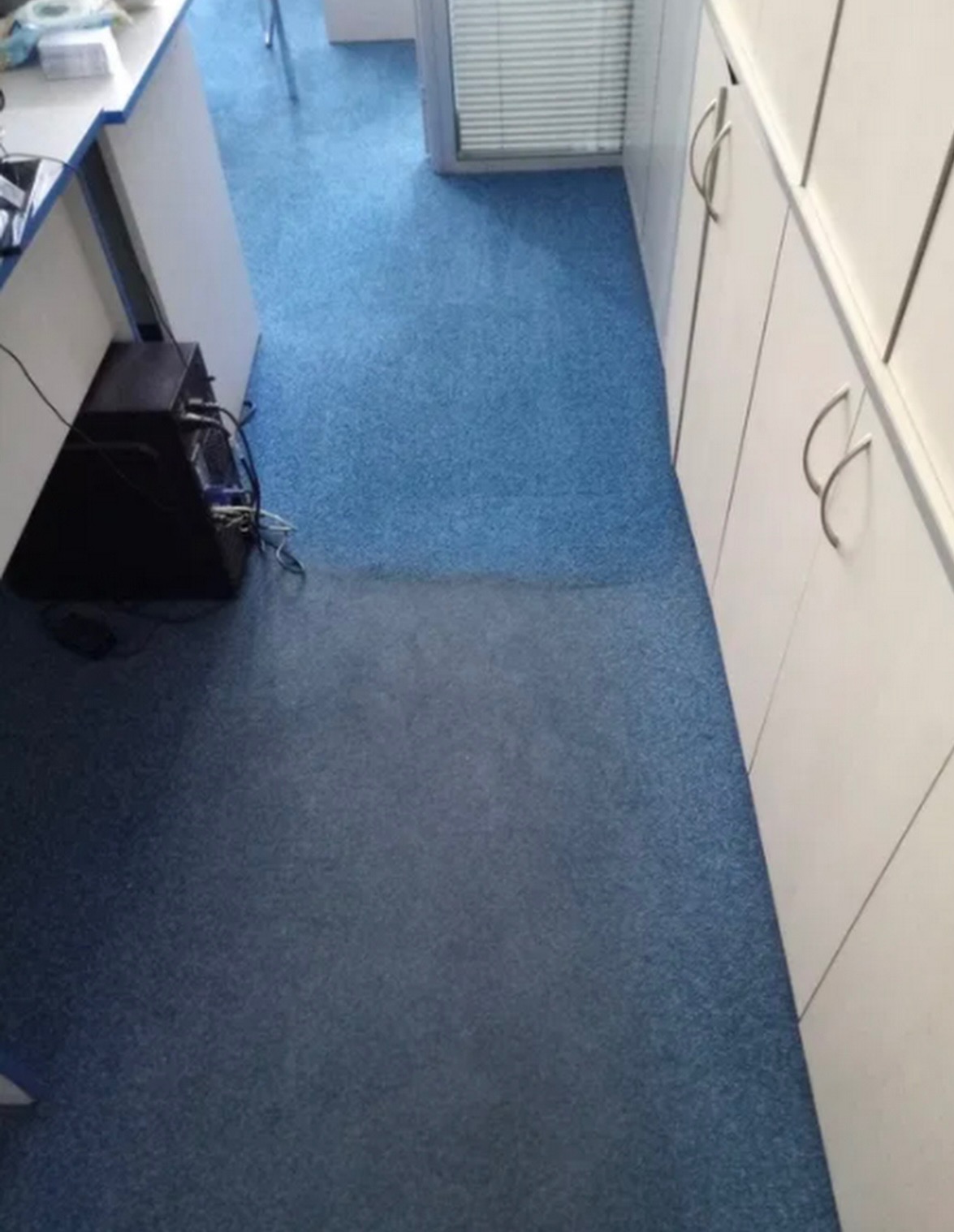 Fragment biura z niebieską wykładziną dywanową, widoczna różnica w odcieniu po czyszczeniu na części powierzchni, przy ścianie białe szafki z metalowymi uchwytami i biurko z komputerem.