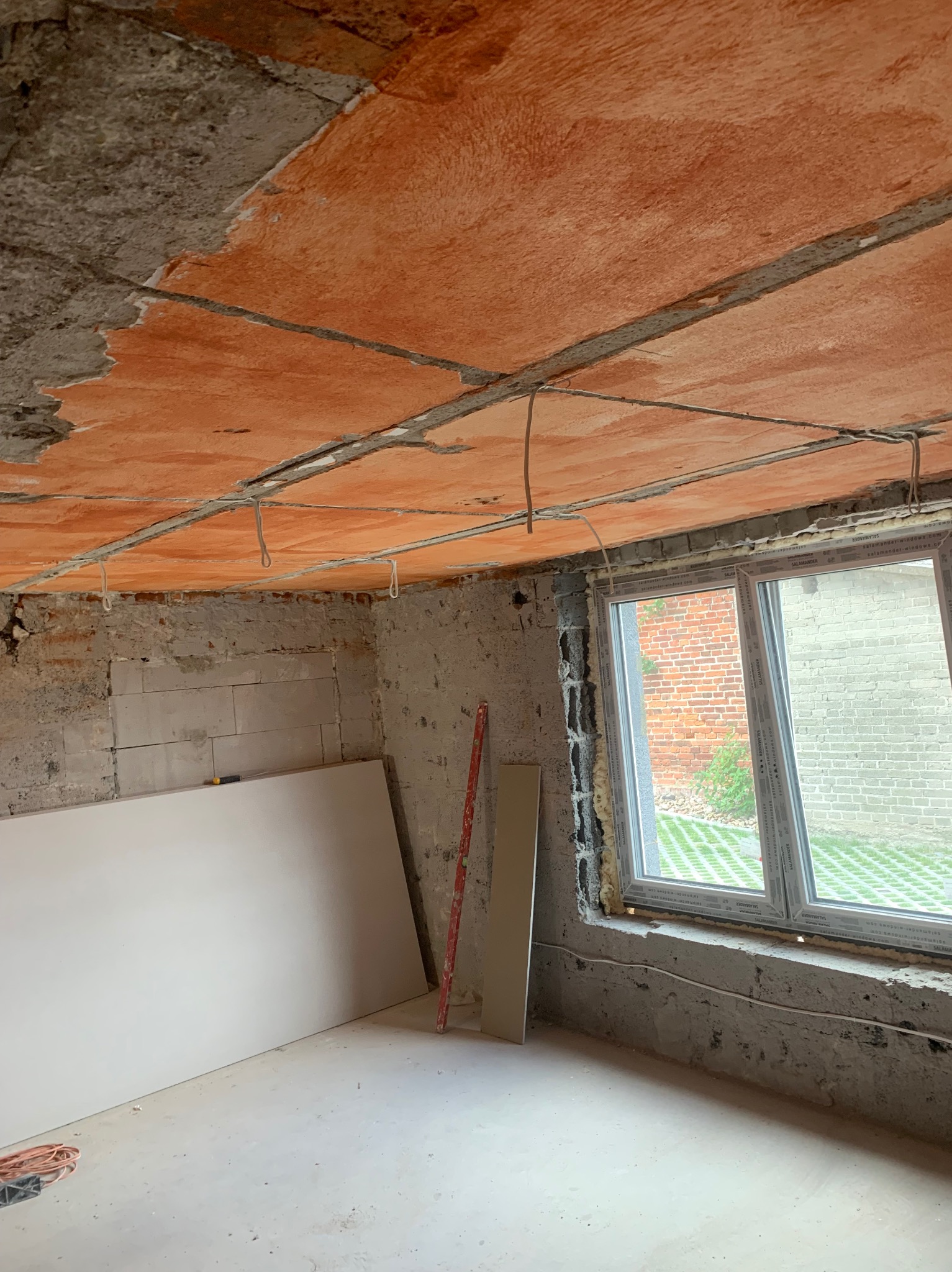 Surowy stan wnętrza: nieotynkowany sufit z widoczną izolacją termiczną, wystające przewody elektryczne, nowe okno PCV, płyty gipsowo-kartonowe oparte o ścianę, poziomica w rogu.