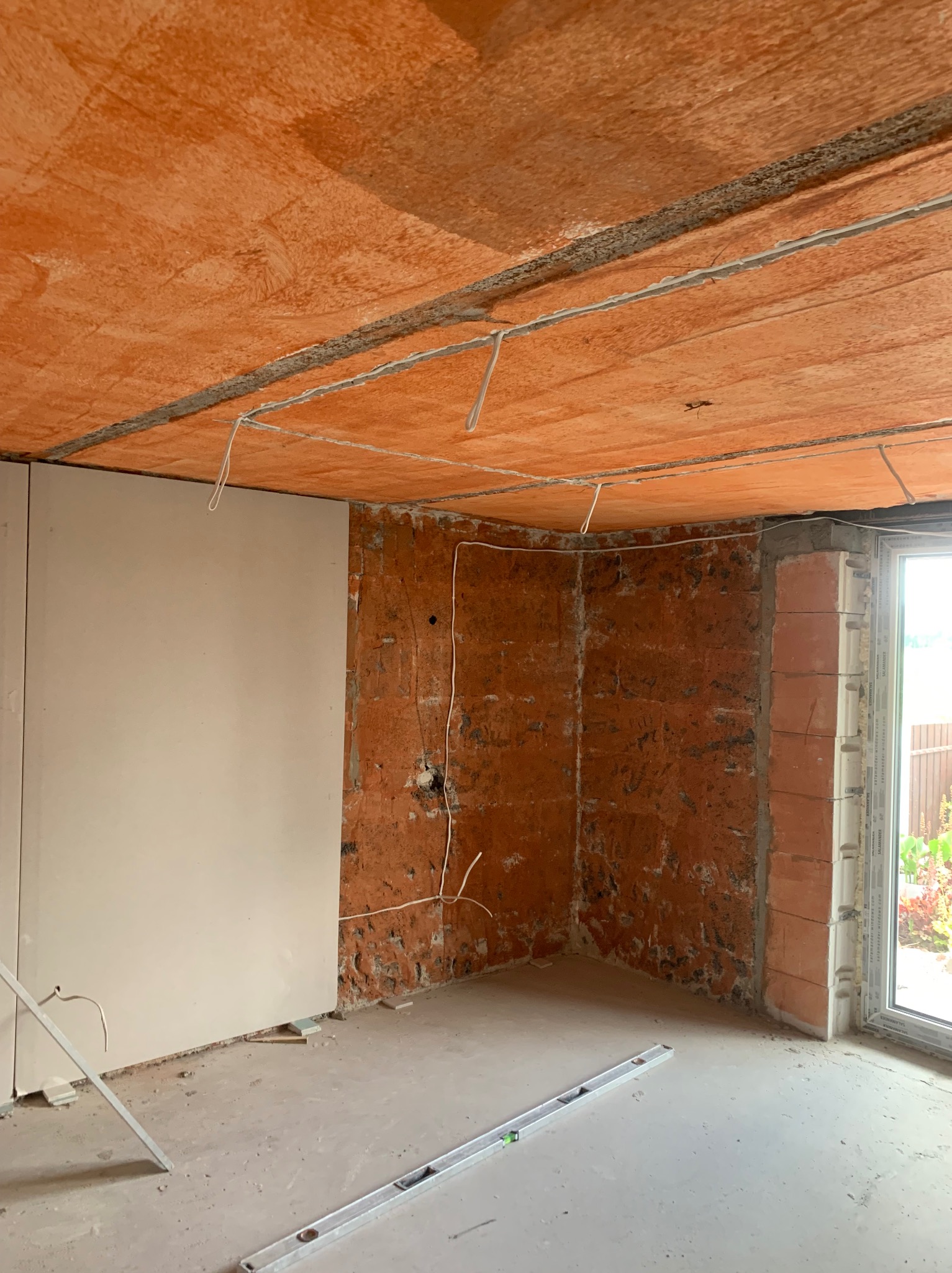 Surowy stan wnętrza z widoczną instalacją elektryczną: przewody wystające z sufitu i ściany z cegły, częściowo otynkowane, poziomica na podłodze, okno w tle.