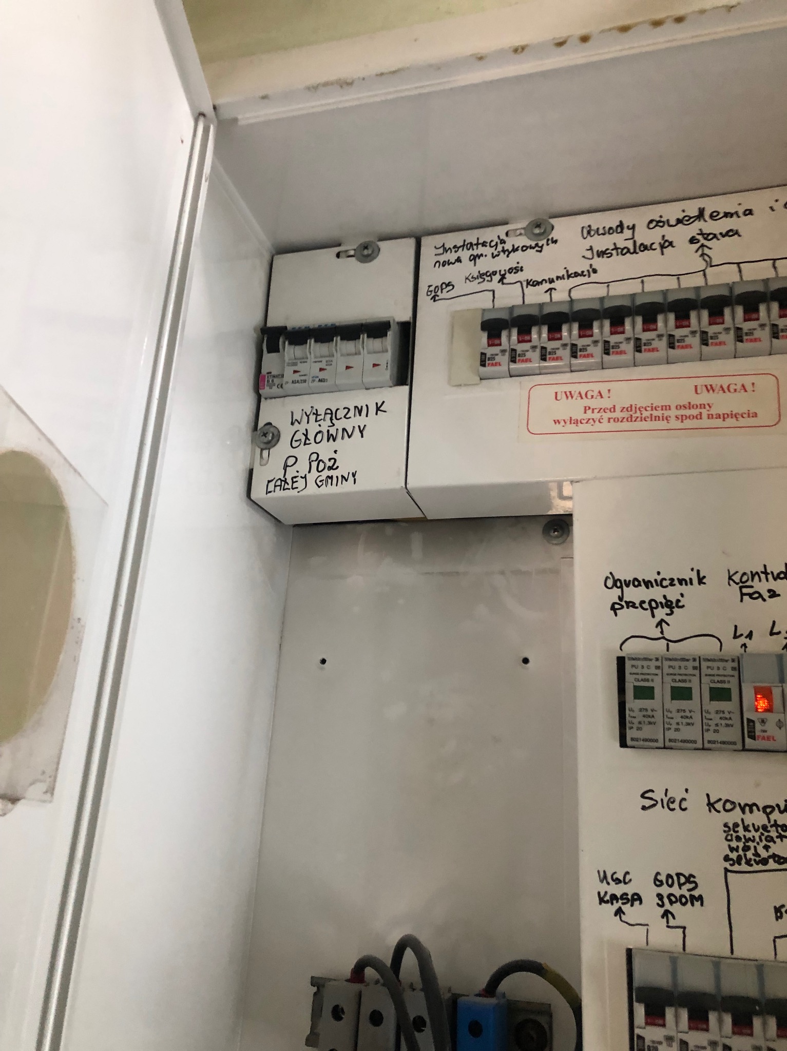 Otwarta skrzynka elektryczna z wyłącznikami, bezpiecznikami i opisami odręcznymi, widoczne okablowanie i zabezpieczenia przeciwprzepięciowe.