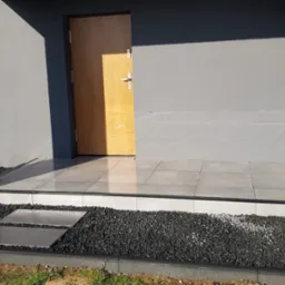 Nowoczesne wejście do domu z szarymi płytkami na podwyższeniu i czarnym kamieniem ozdobnym, widoczne dwa płyty chodnikowe prowadzące do schodów.