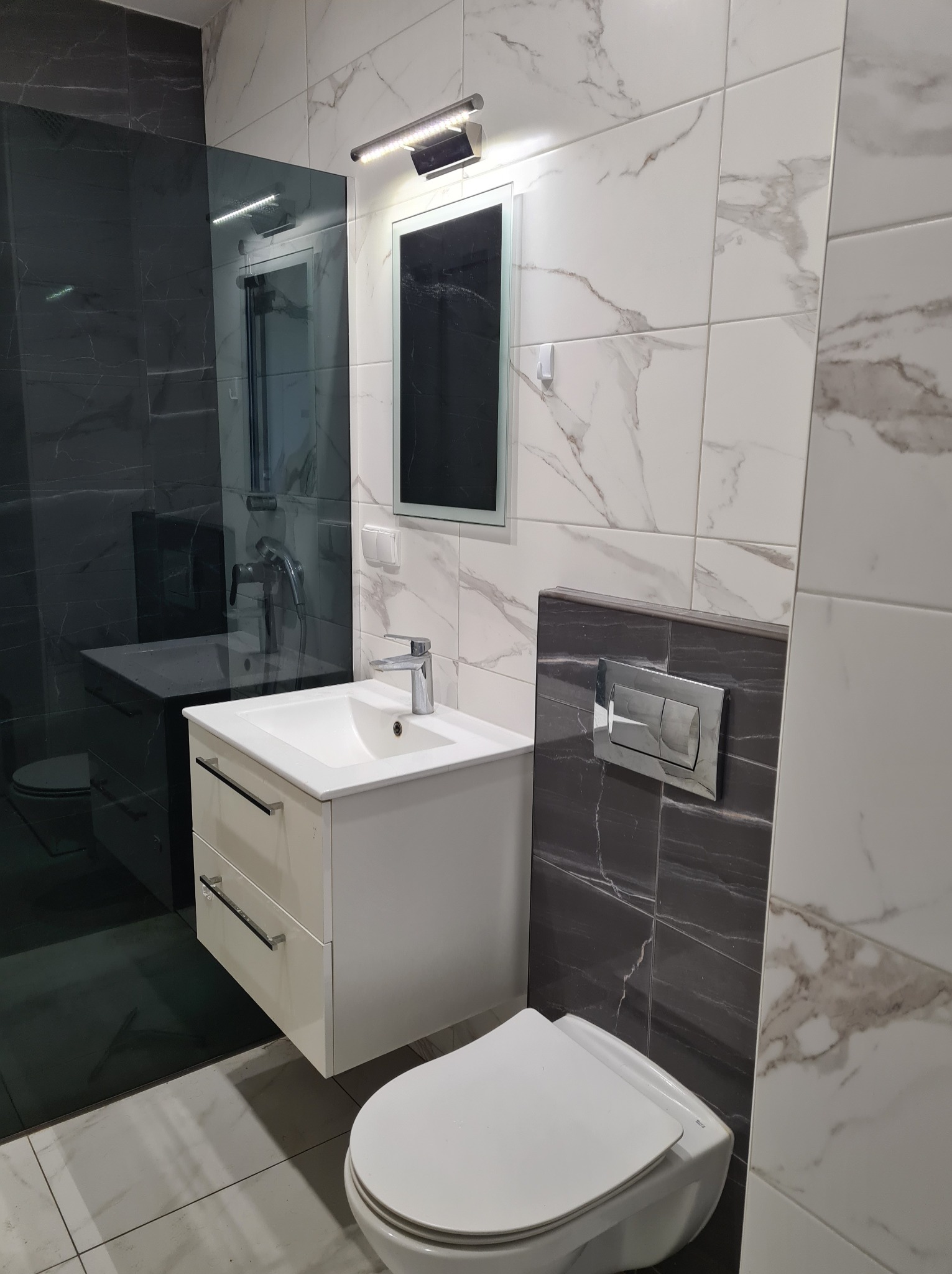 Nowoczesna łazienka z białymi i szarymi płytkami imitującymi marmur, wisząca szafka z umywalką, toaleta i kabina prysznicowa ze szkła.