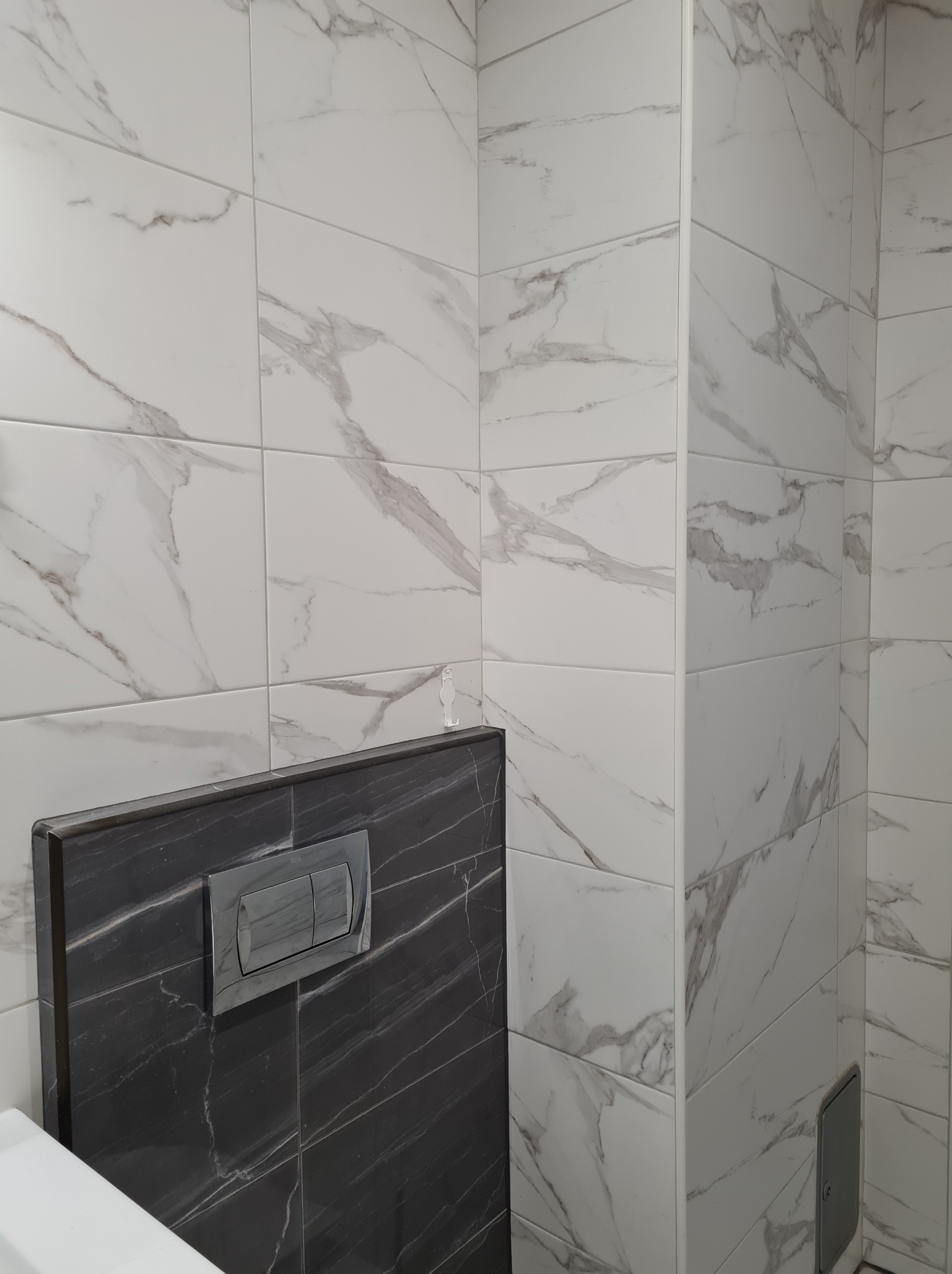 Wykończona toaleta z białymi płytkami imitującymi marmur na ścianach i zabudową stelaża WC z czarnych płytek z szarym przyciskiem spłukującym.