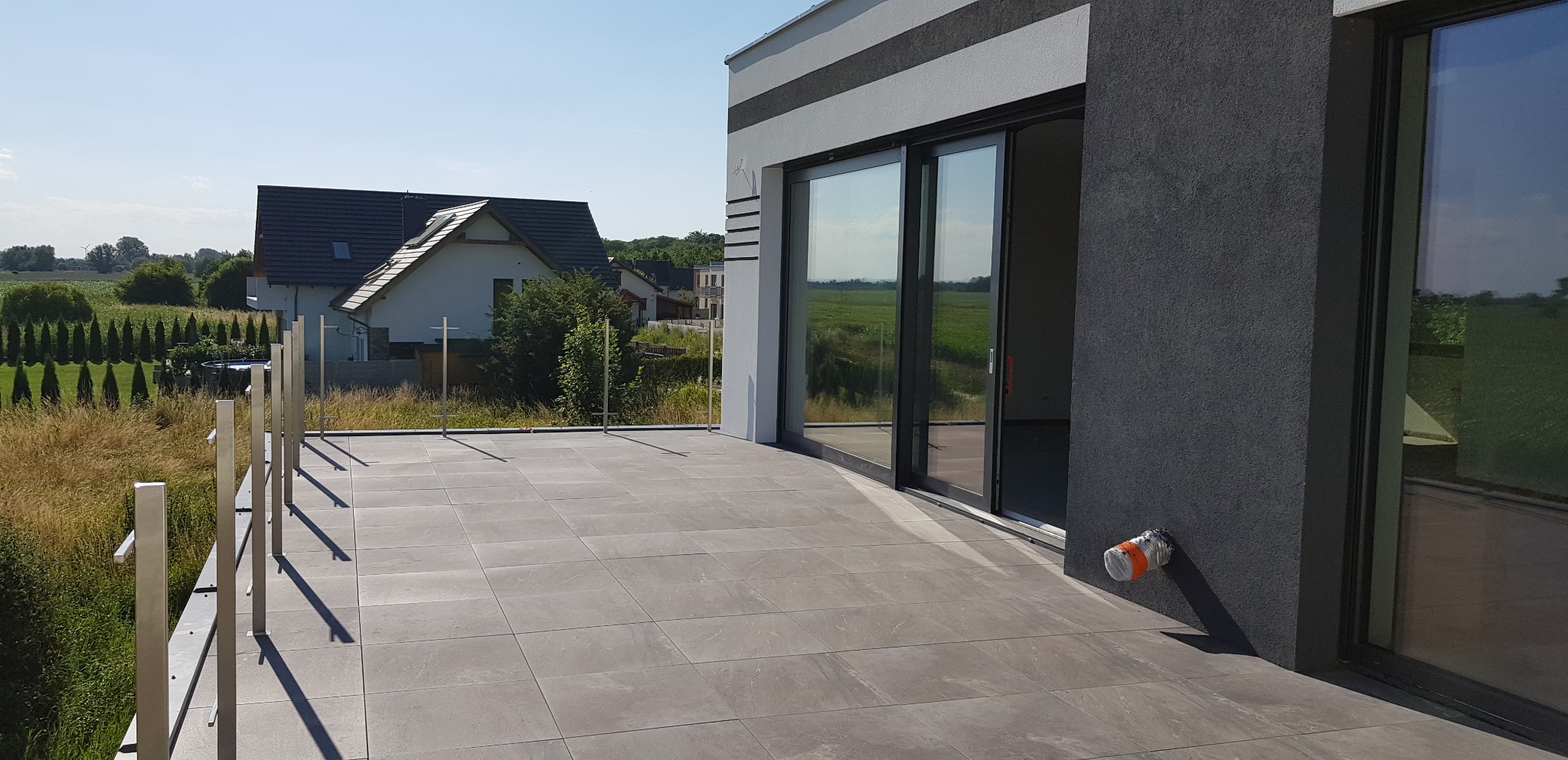 Nowoczesny taras wyłożony szarymi płytkami, ze stalową balustradą, widok na okoliczne domy i pola w słoneczny dzień.