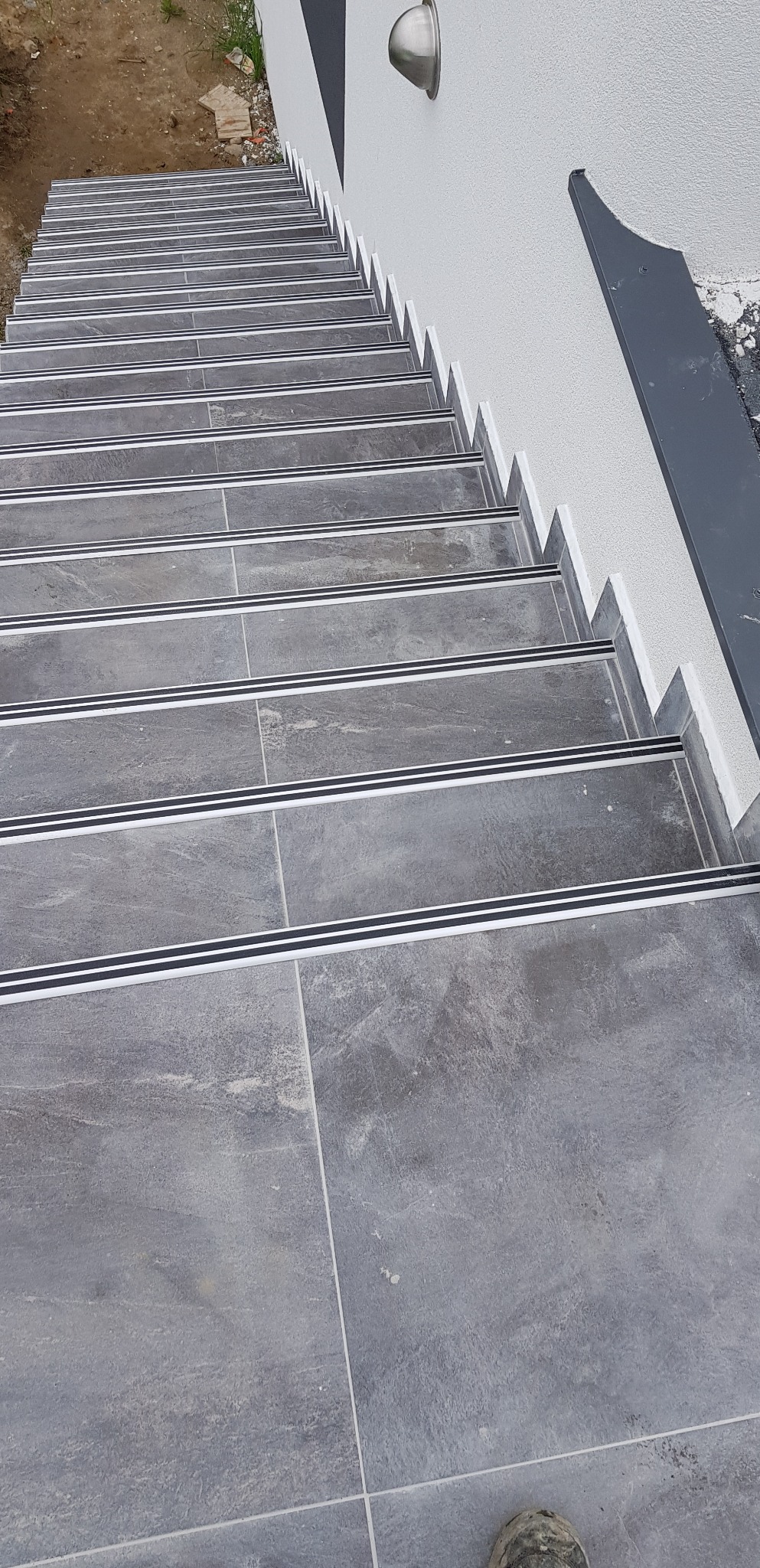 Zewnętrzne schody wyłożone szarymi płytkami z antypoślizgowymi listwami, widok z góry w kierunku zejścia, fragment buta roboczego w dolnym rogu.