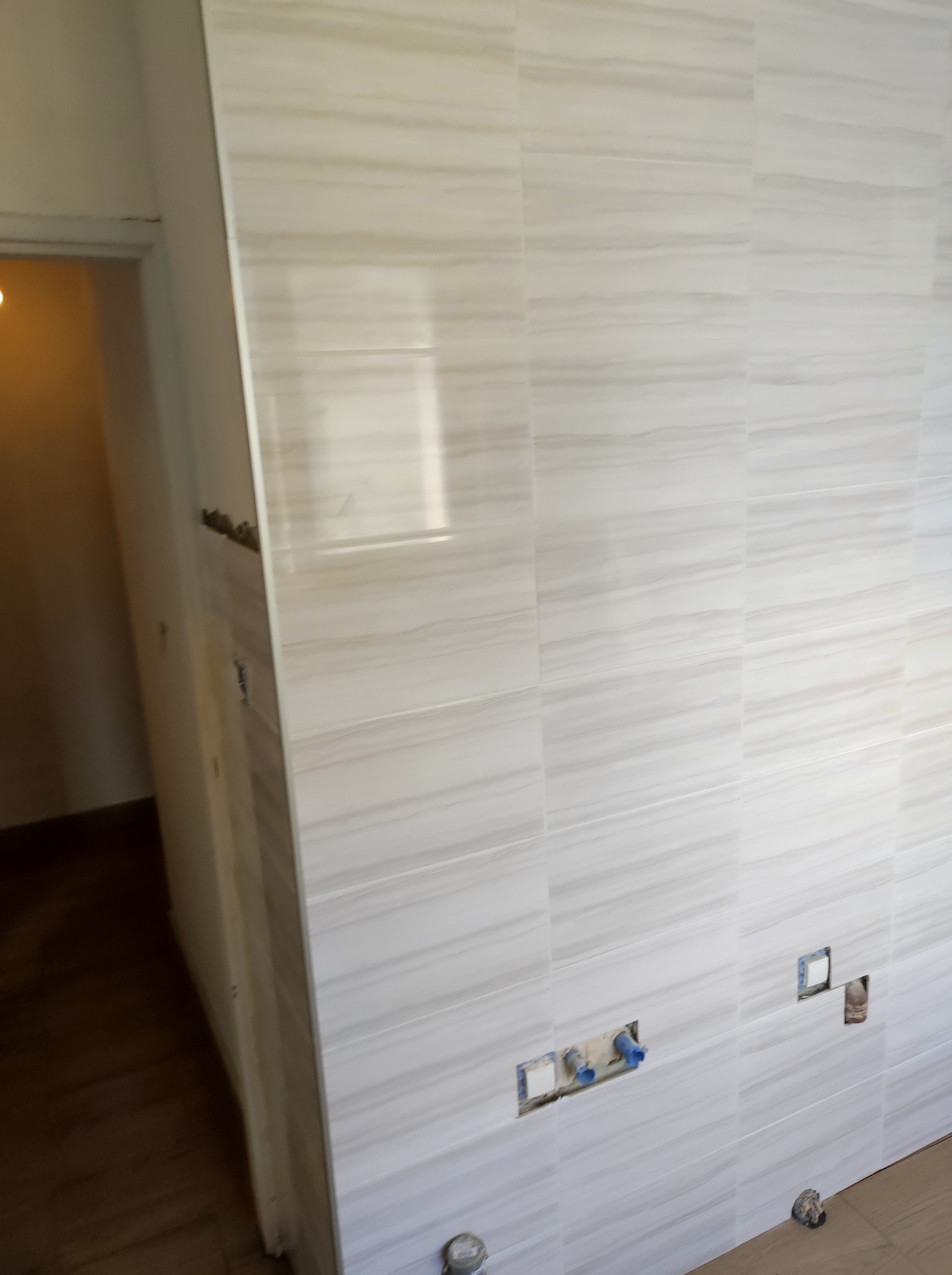 Ściana w łazience wyłożona płytkami imitującymi marmur, z widocznymi otworami na instalacje wodno-kanalizacyjne, częściowo wykończona.