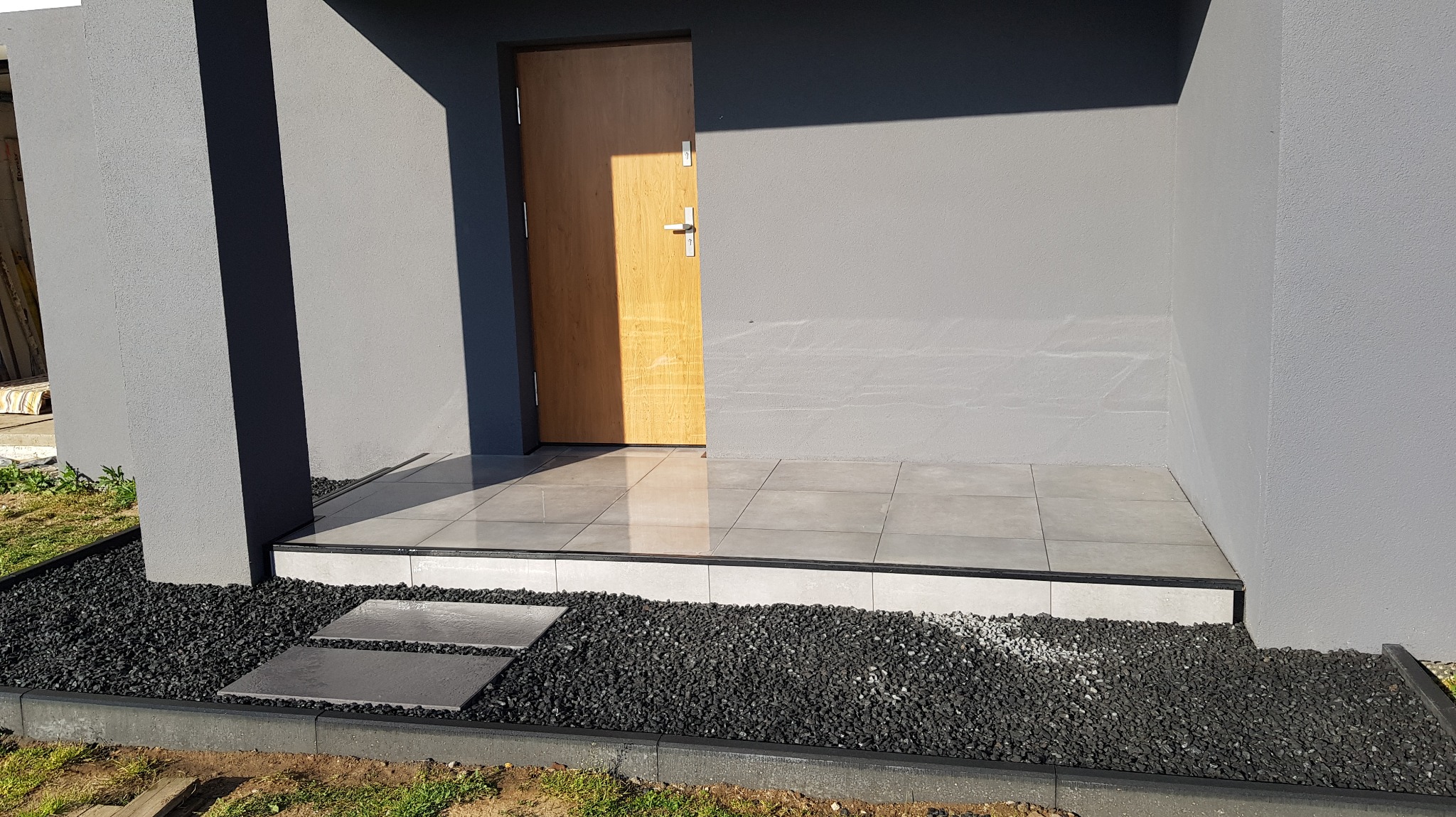 Nowoczesne wejście do domu z szarymi płytkami na podwyższeniu i czarnym kamieniem ozdobnym, widoczne dwa płyty chodnikowe prowadzące do schodów.