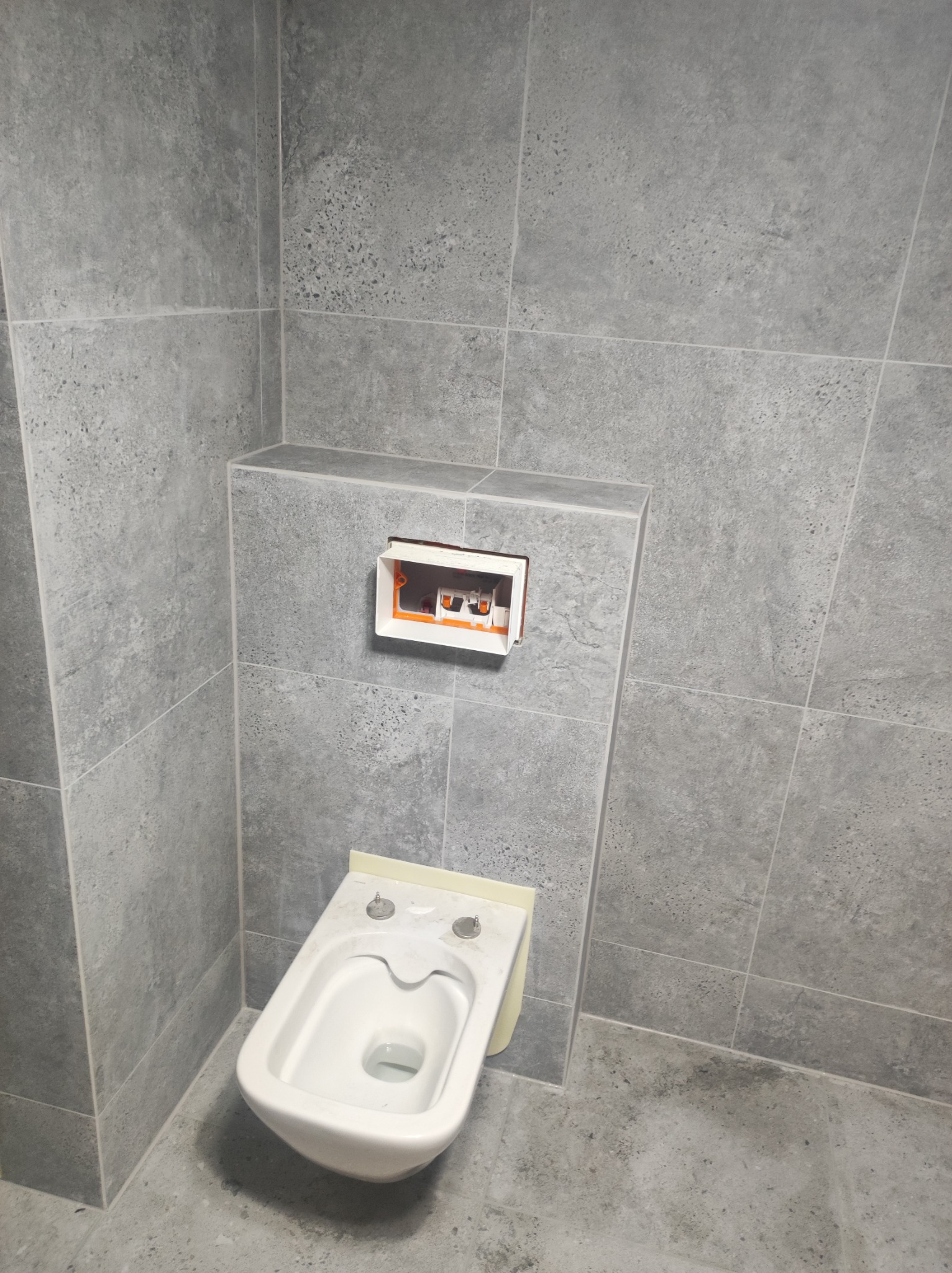 Wisząca miska WC w trakcie montażu, obudowana szarą płytką imitującą beton, widoczna puszka instalacyjna podtynkowego systemu spłukiwania.