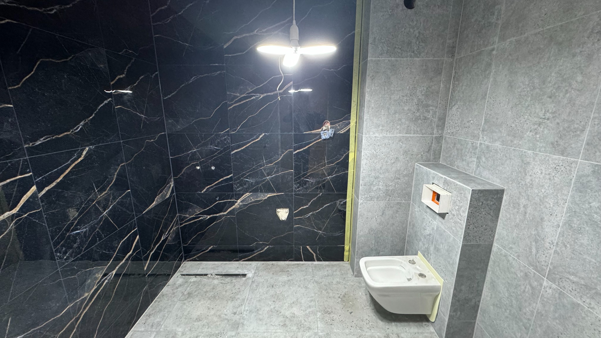 Wnętrze łazienki w trakcie wykańczania, z czarnymi płytkami imitującymi marmur na ścianie prysznicowej i szarymi płytkami na pozostałych ścianach, widoczne niedokończone podłączenie bidetu...