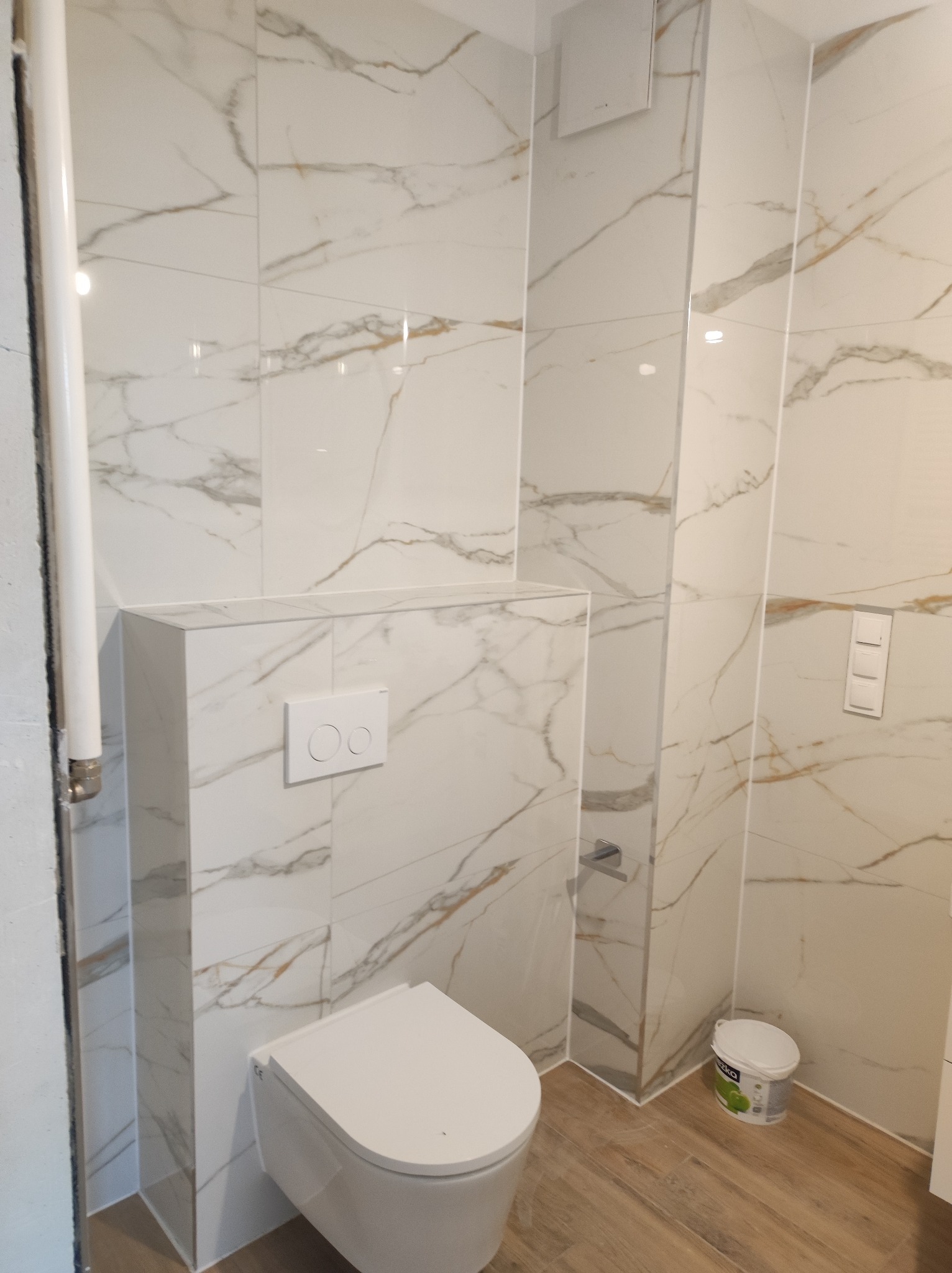 Zabudowana toaleta podwieszana z białymi płytkami imitującymi marmur z delikatnymi złotymi żyłkami, drewnopodobna podłoga, biały przycisk spłuczki i widoczne wiaderko po farbie.